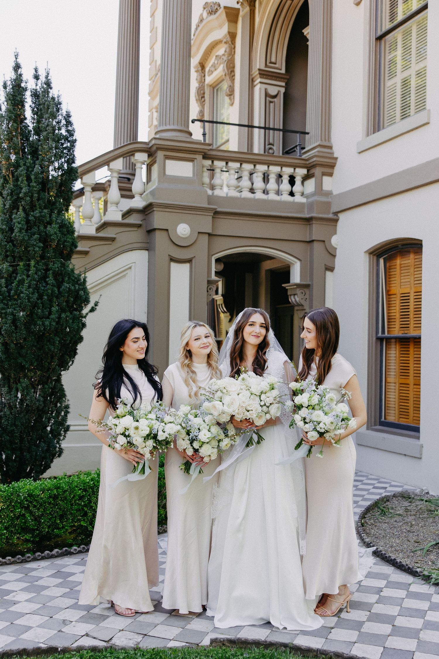 Wedding party in cream-colored gowns holding white floral bouquets pose outside elegant mansion with classical architecture.