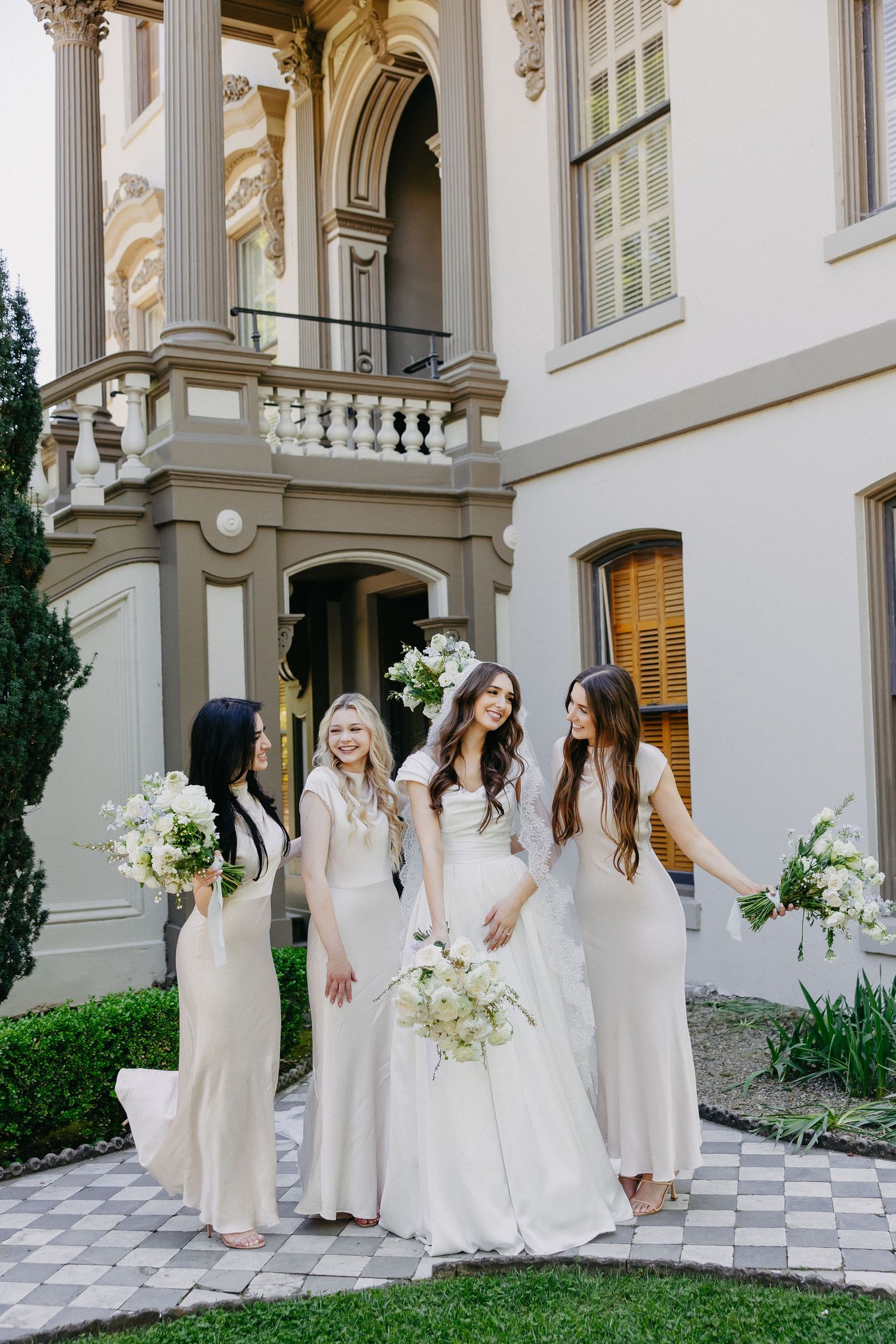 Bridal party shares joyful moment on checkered courtyard of historic building with ornate columns.