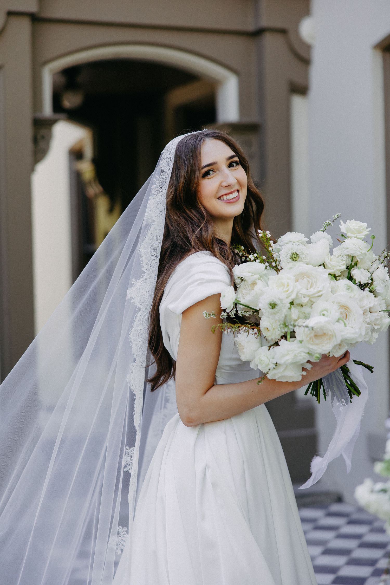 Bride in classic white dress holds luxurious white bouquet while smiling beside elegant mansion.