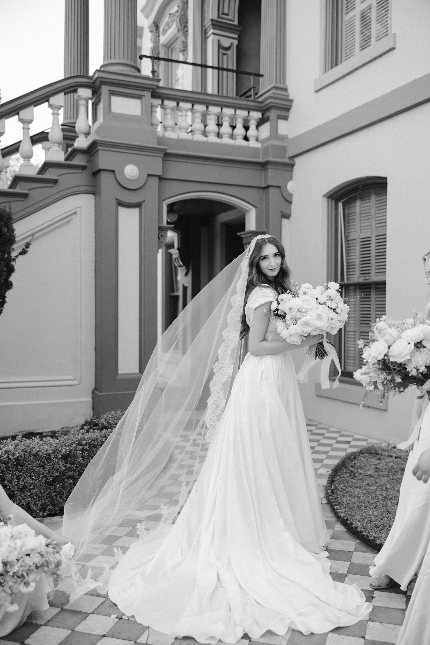 Black and white portrait of bride with flowing veil standing on checkered courtyard.