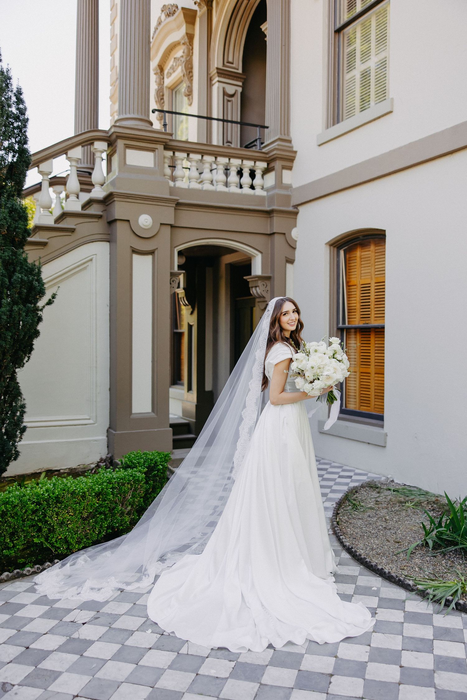 Elegant bride with cathedral veil poses with white bouquet against classical architecture backdrop.