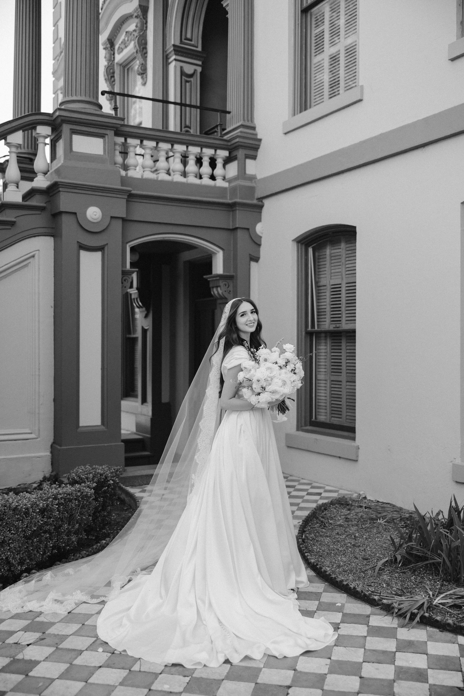Black and white bridal portrait showcasing long train and veil against historic building facade.