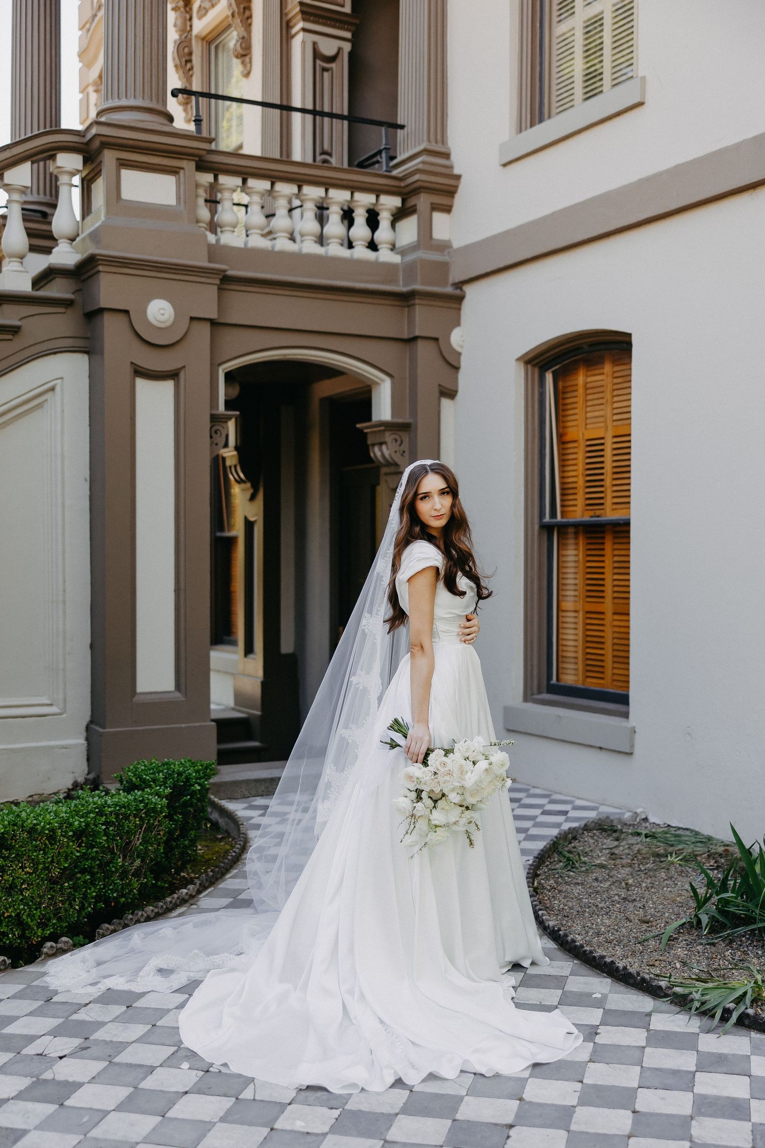 Bride in flowing gown turns gracefully on checkered path beside ornate mansion entrance.