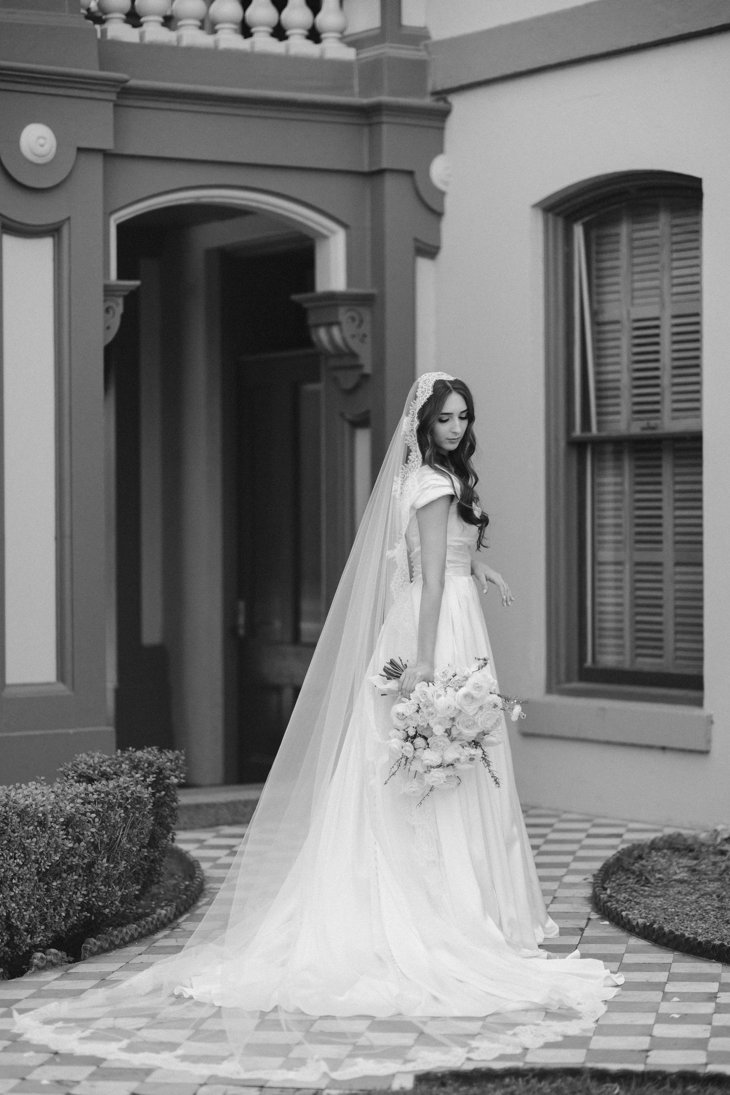 Monochrome capture of bride in full-length gown standing beside historic building entrance.