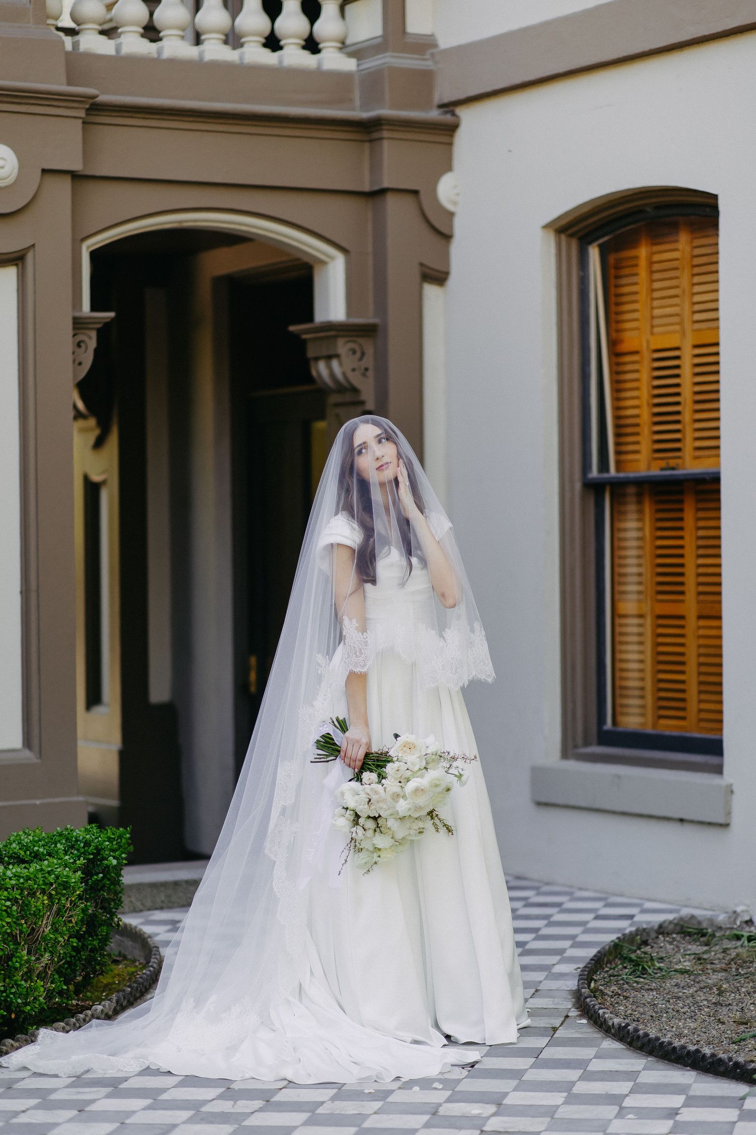 Ethereal bridal portrait with flowing veil and white bouquet against classical architecture.
