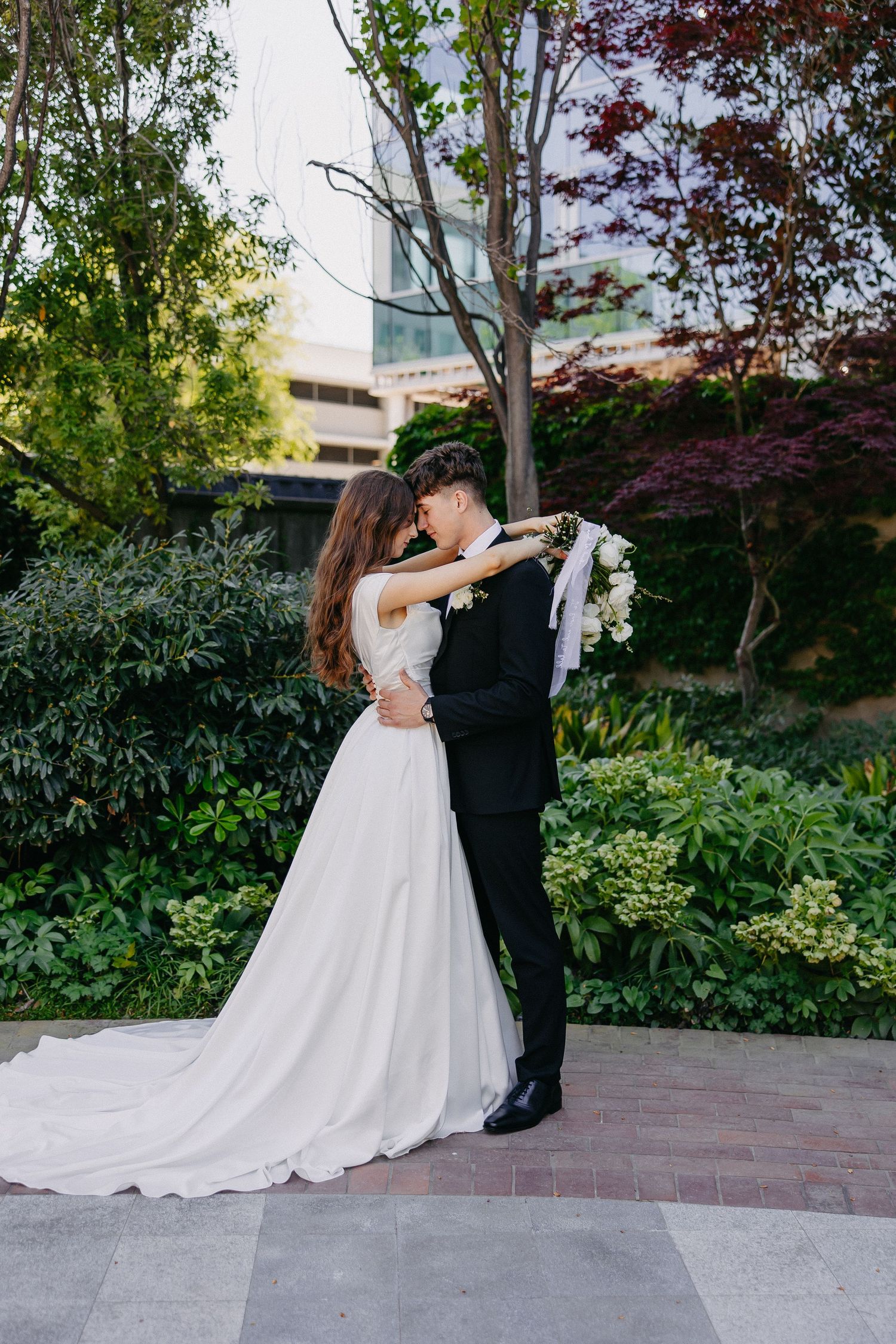 A couple embraces during their wedding photoshoot in a lush garden setting with a flowing white dress and classic tuxedo.