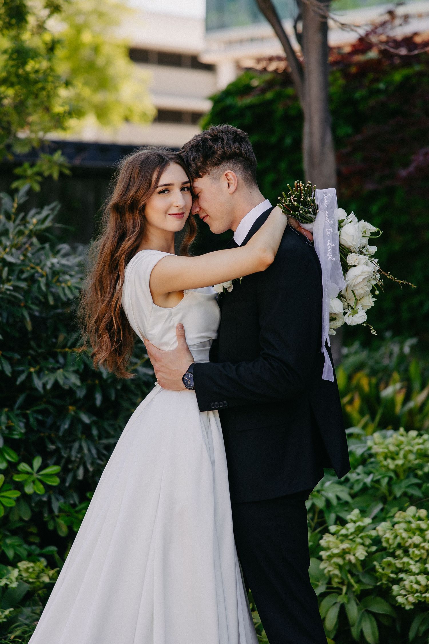 Newlyweds share an intimate moment in a garden with the bride wearing a flowy white gown and groom in black suit.