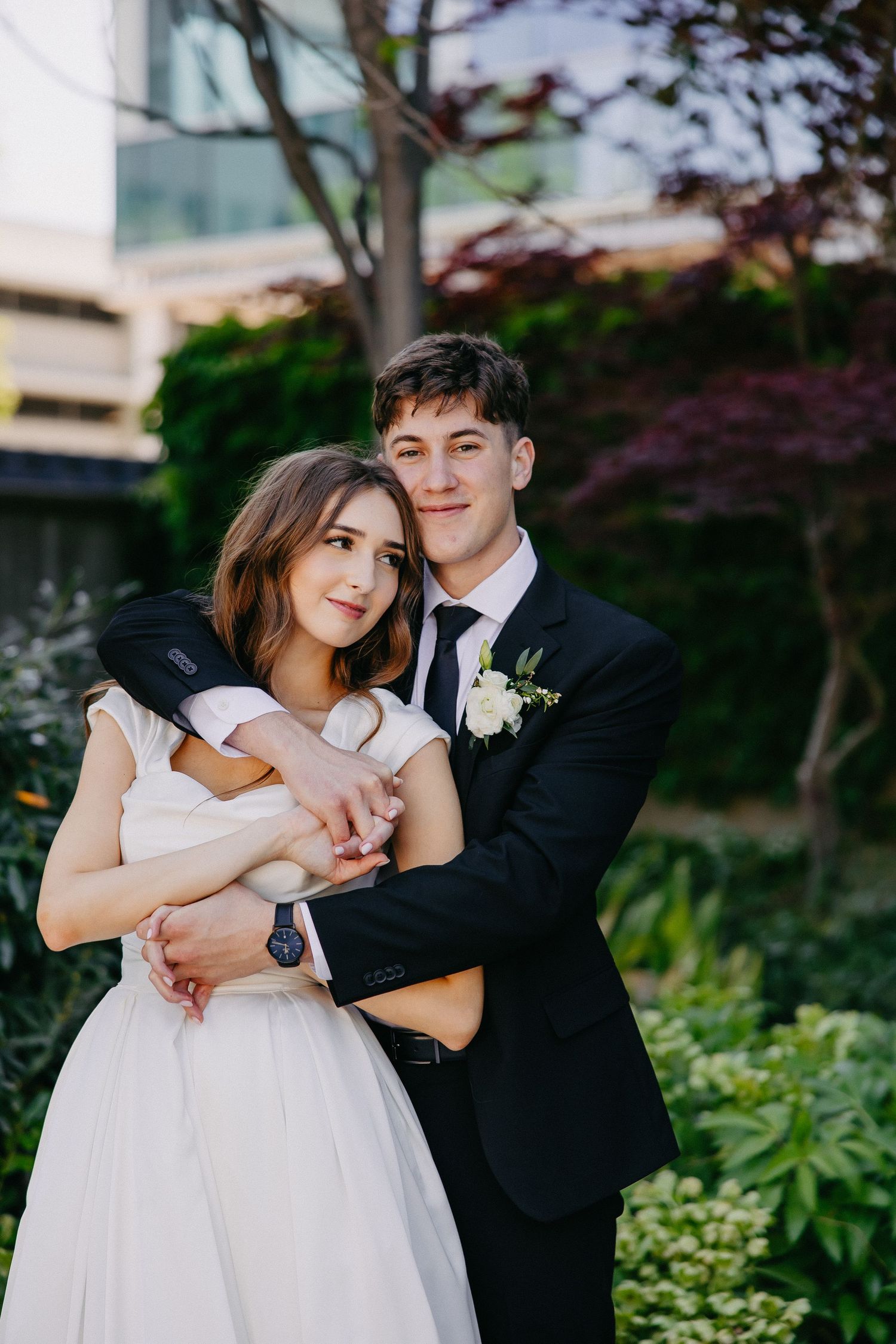 Wedding couple poses in a garden with modern architecture backdrop, sharing a playful moment together.