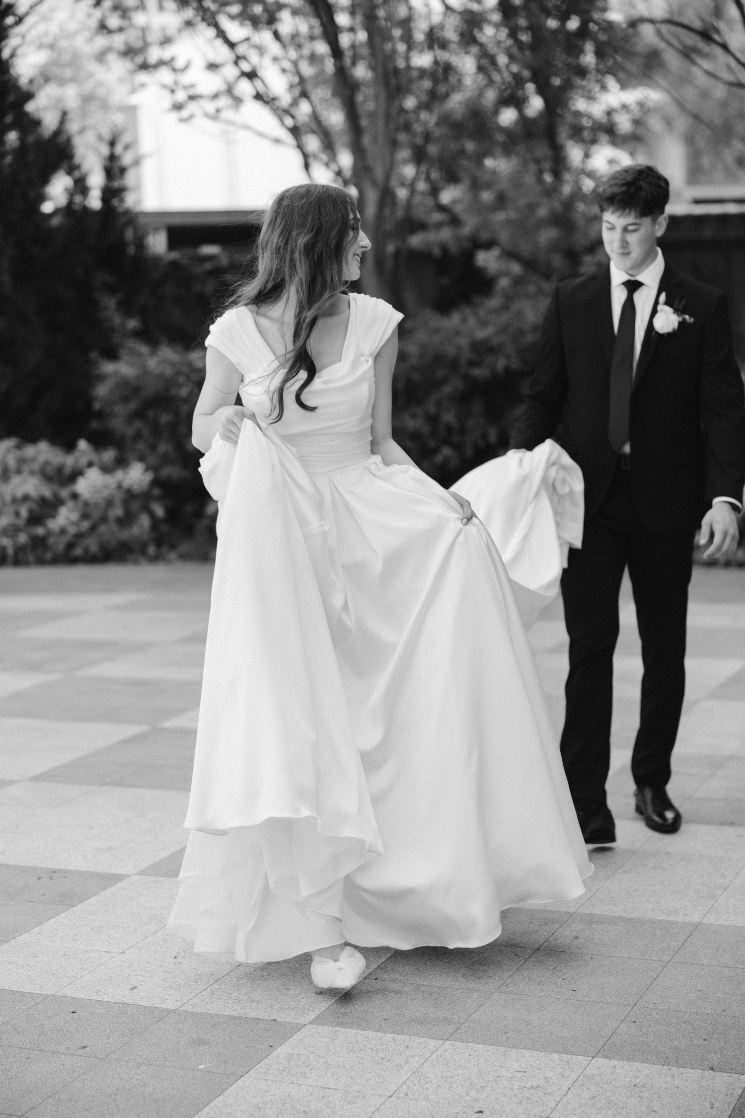 Black and white photo of bride twirling her dress while groom watches on stone patio.