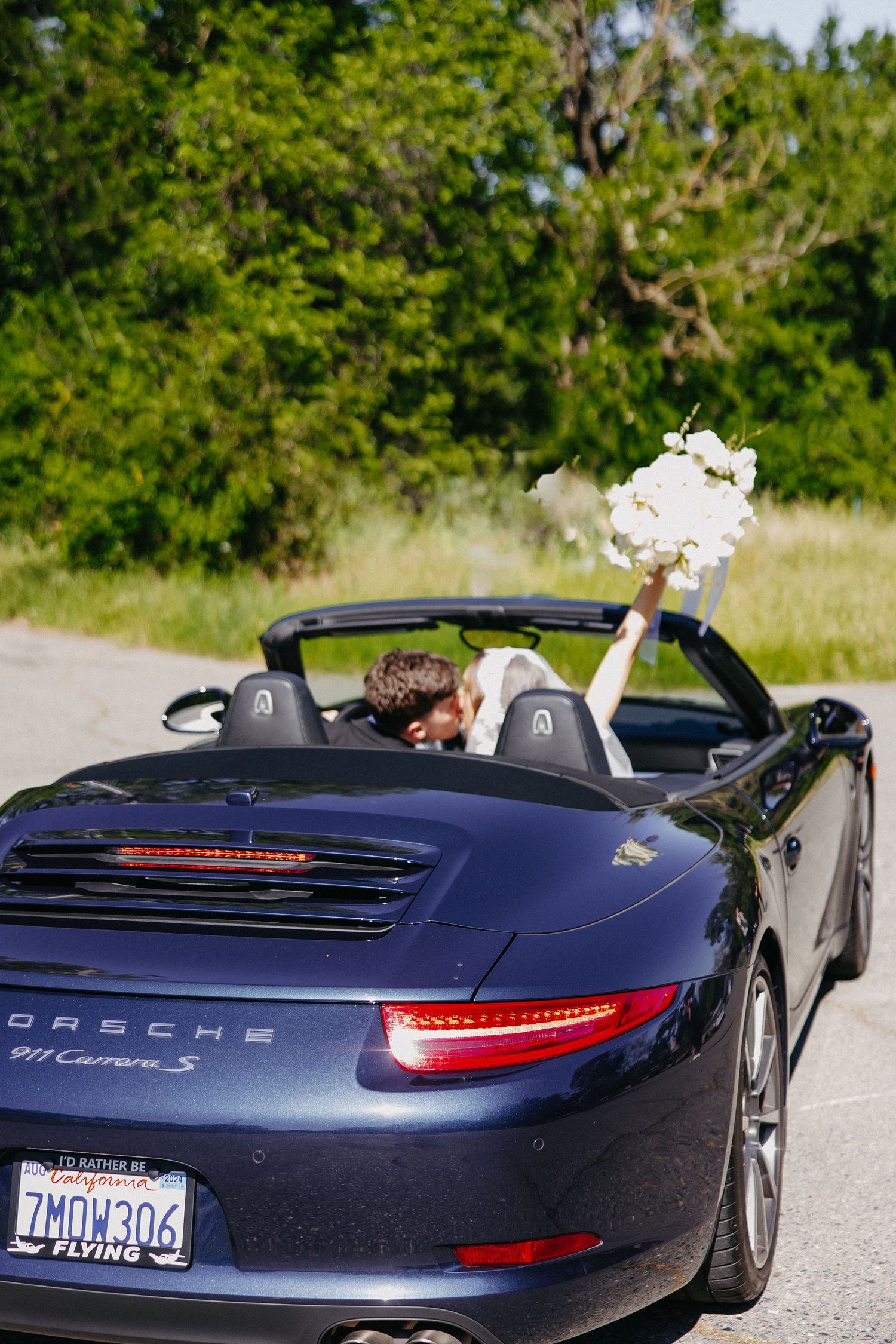 Couple celebrates wedding day in a navy blue Porsche 911 convertible with white bouquet on a sunny day.