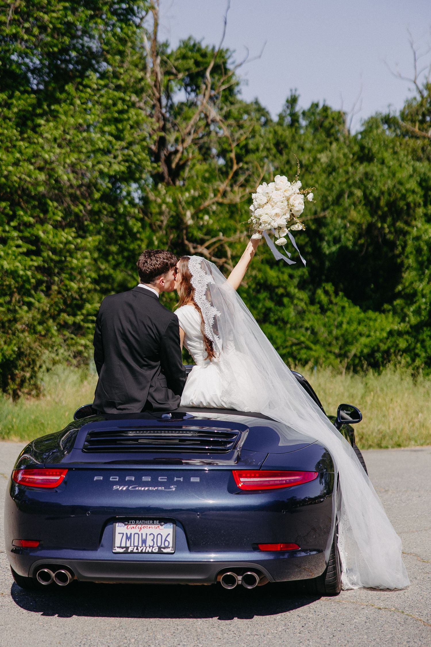 Newlyweds sit on back of dark blue Porsche 911 with bride holding white bouquet against green backdrop.