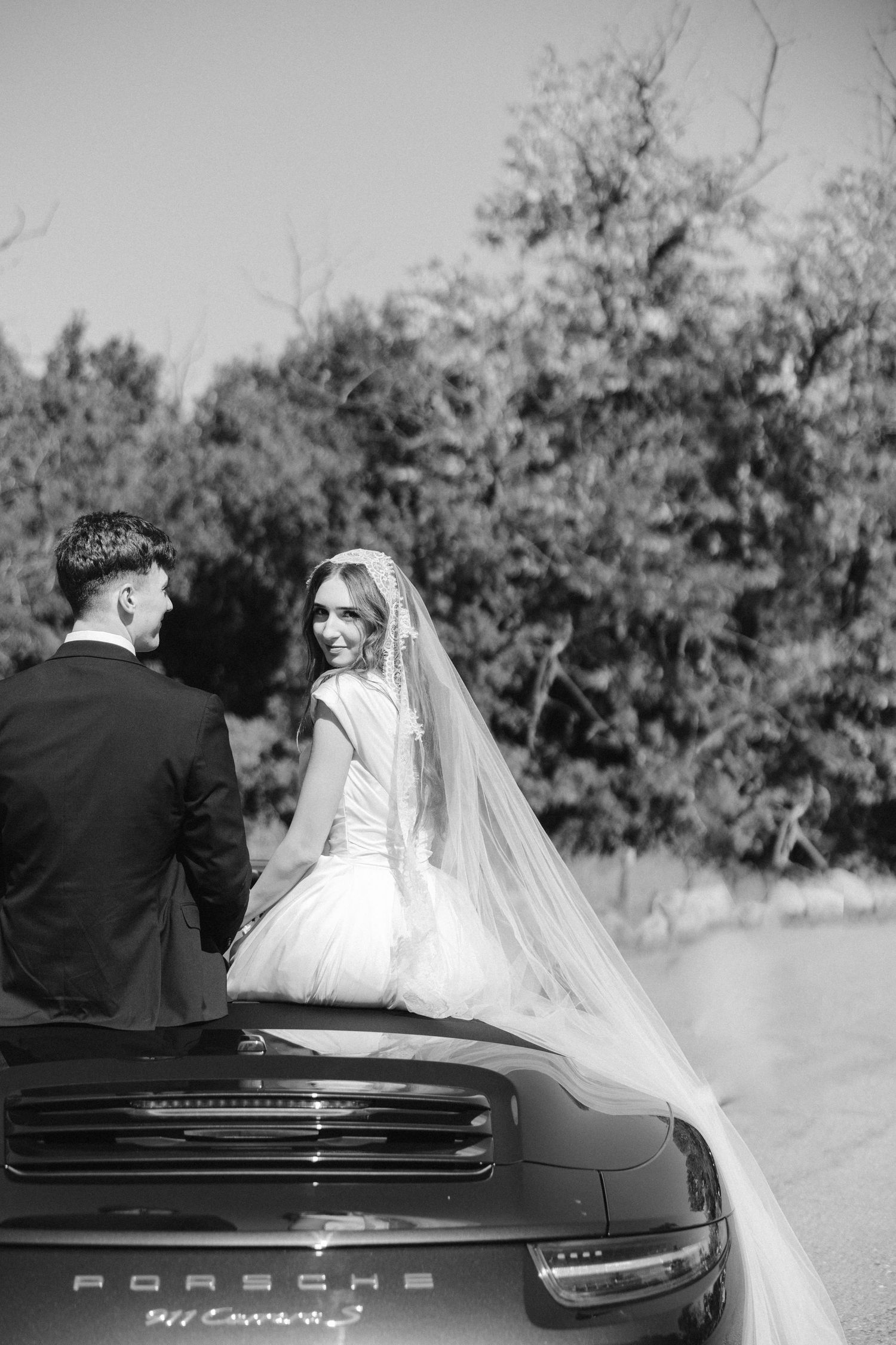 Black and white photo of wedding couple sitting on Porsche convertible trunk.