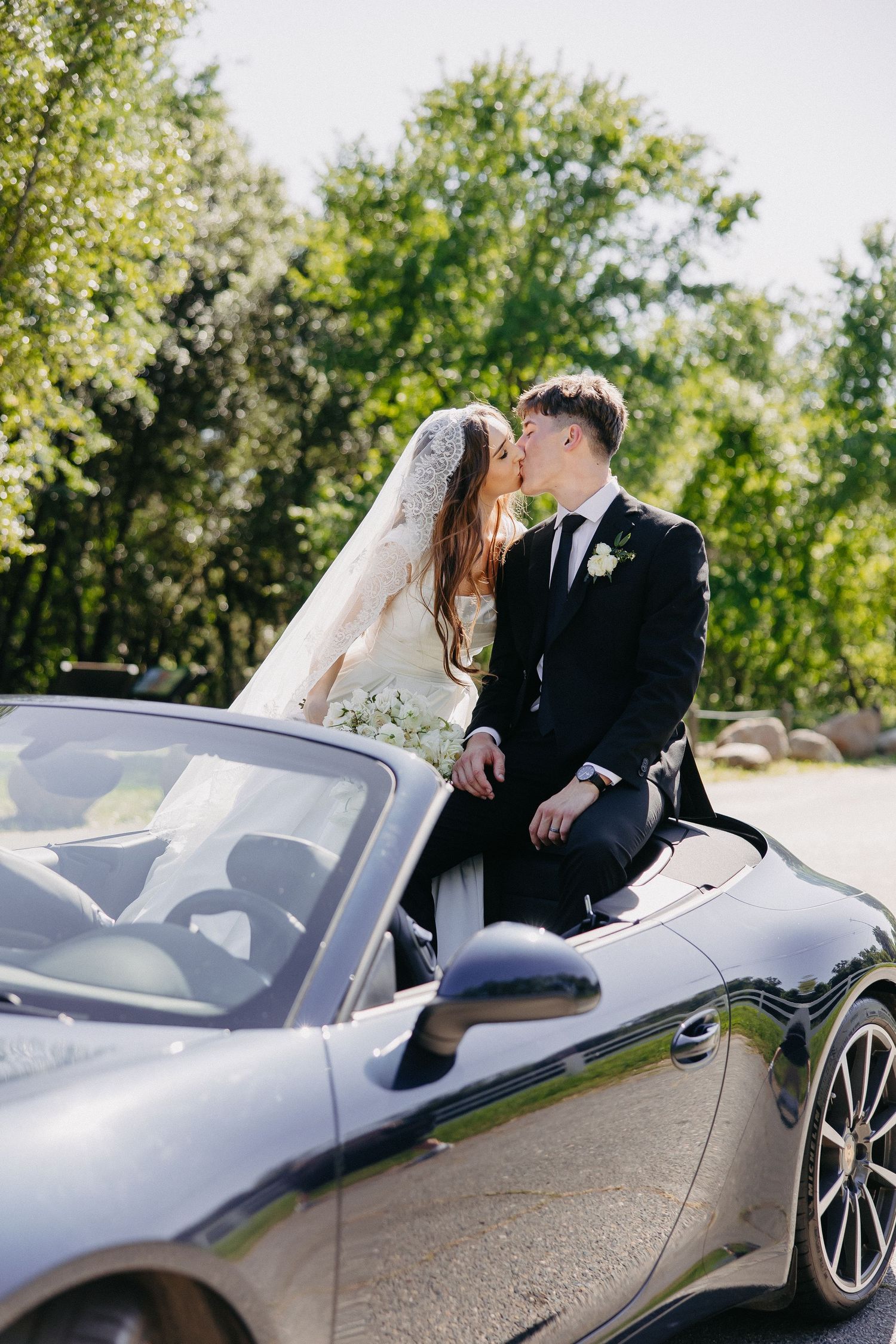 Romantic wedding moment captured with couple on navy Porsche convertible surrounded by trees.