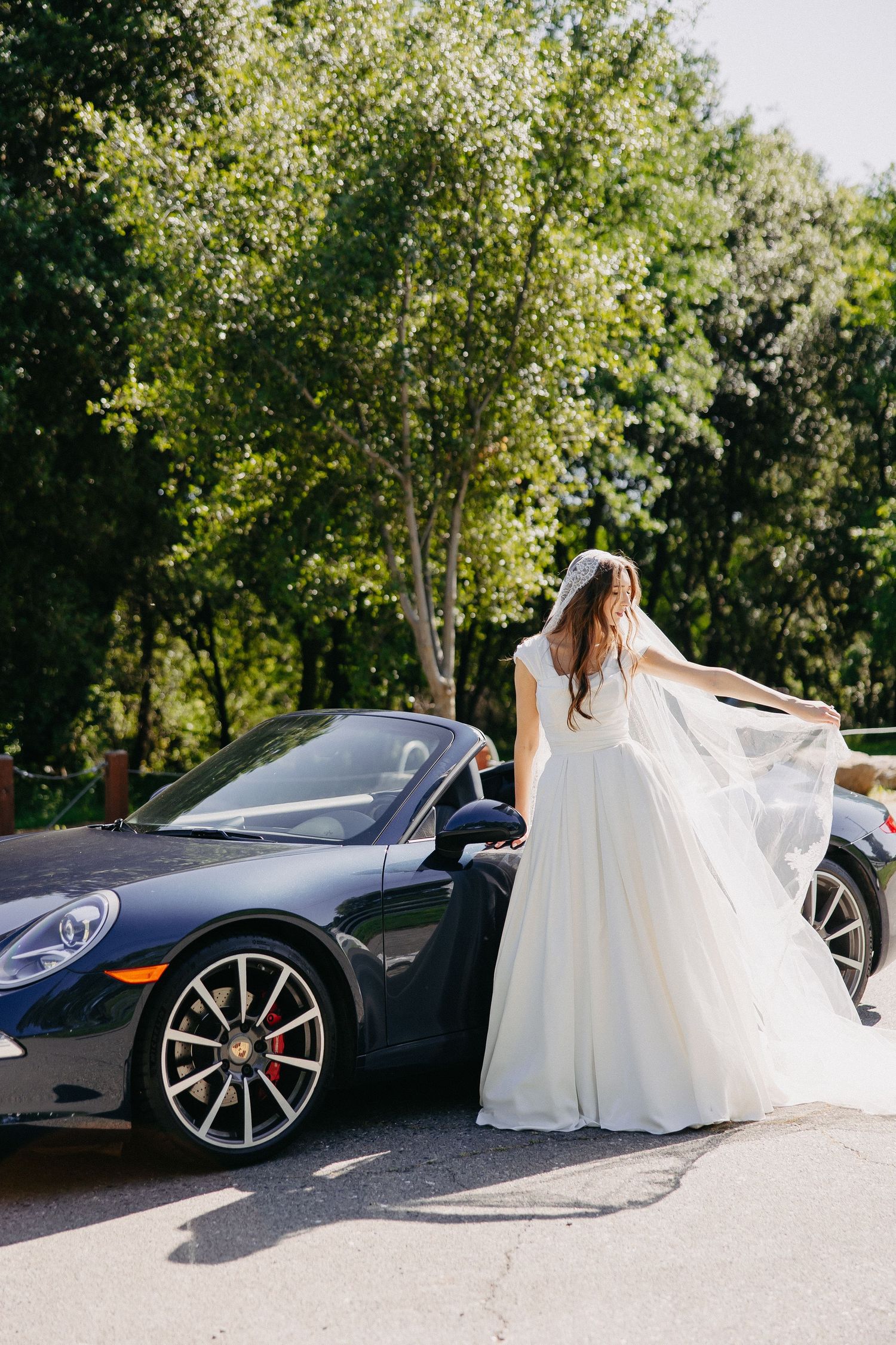 Bride in flowing white gown poses beside dark Porsche 911 convertible on sunny day.
