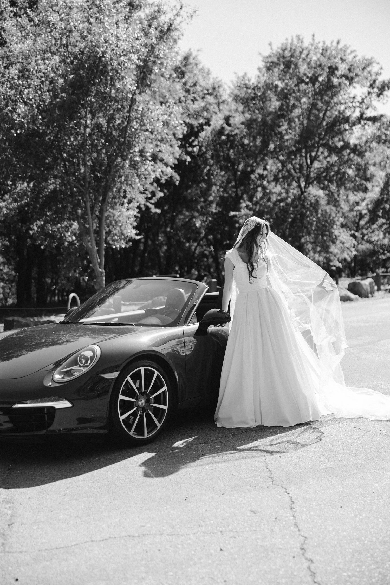 Black and white portrait of bride in flowing dress standing by Porsche sports car.