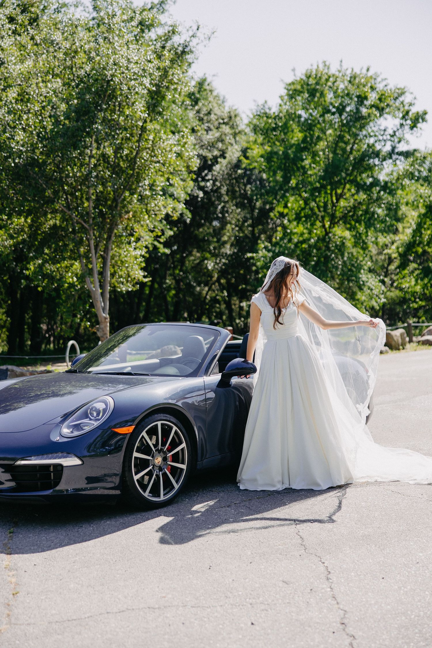 Bride's dress and veil flow in breeze as she stands beside luxury Porsche convertible.