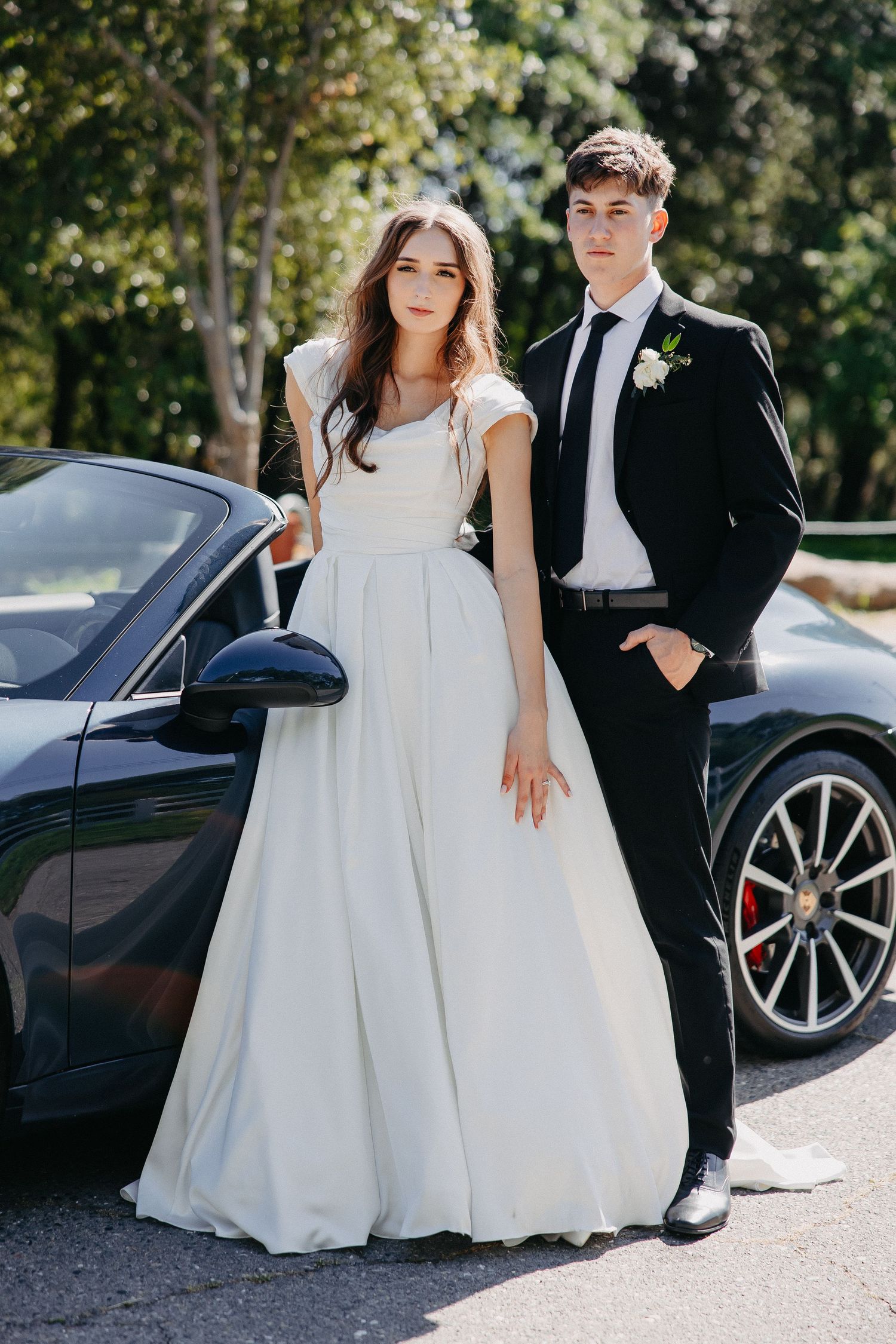 Elegant wedding couple poses beside dark blue Porsche sports car on sunny afternoon.