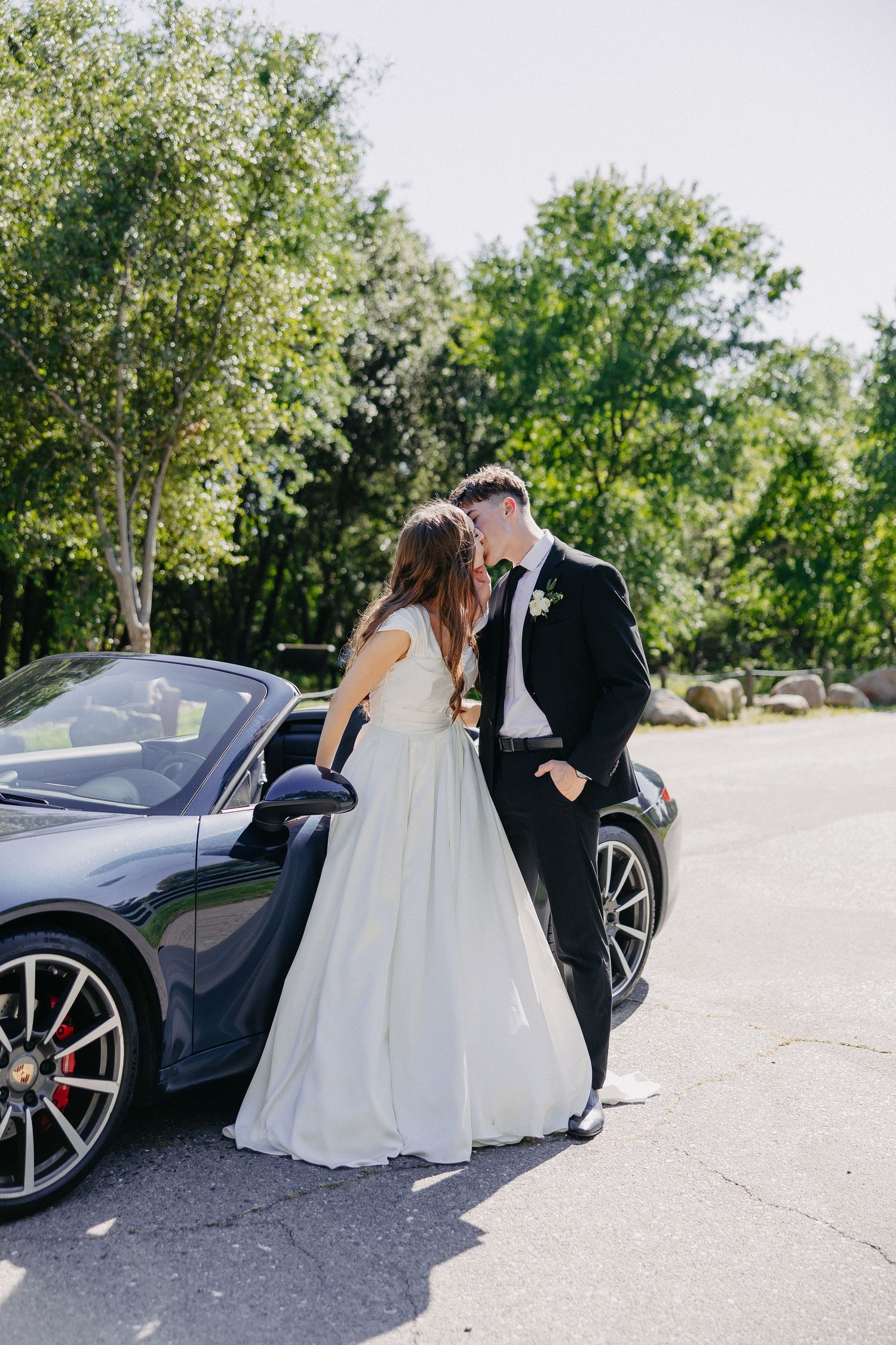 A couple in wedding attire poses next to a dark luxury sports car on a sunny day.