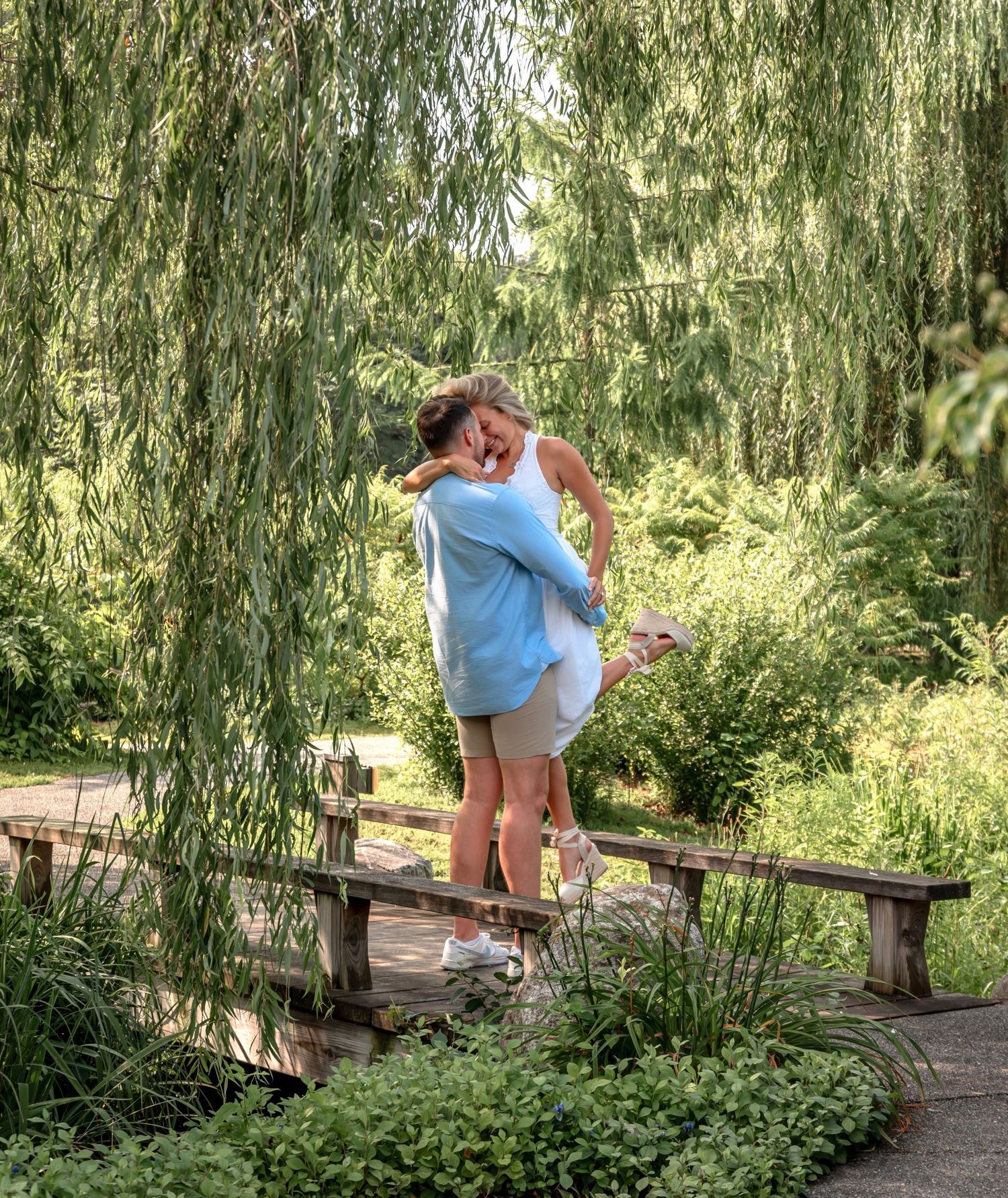 A couple in white share an intimate moment outdoors with sunlit trees in the background.
