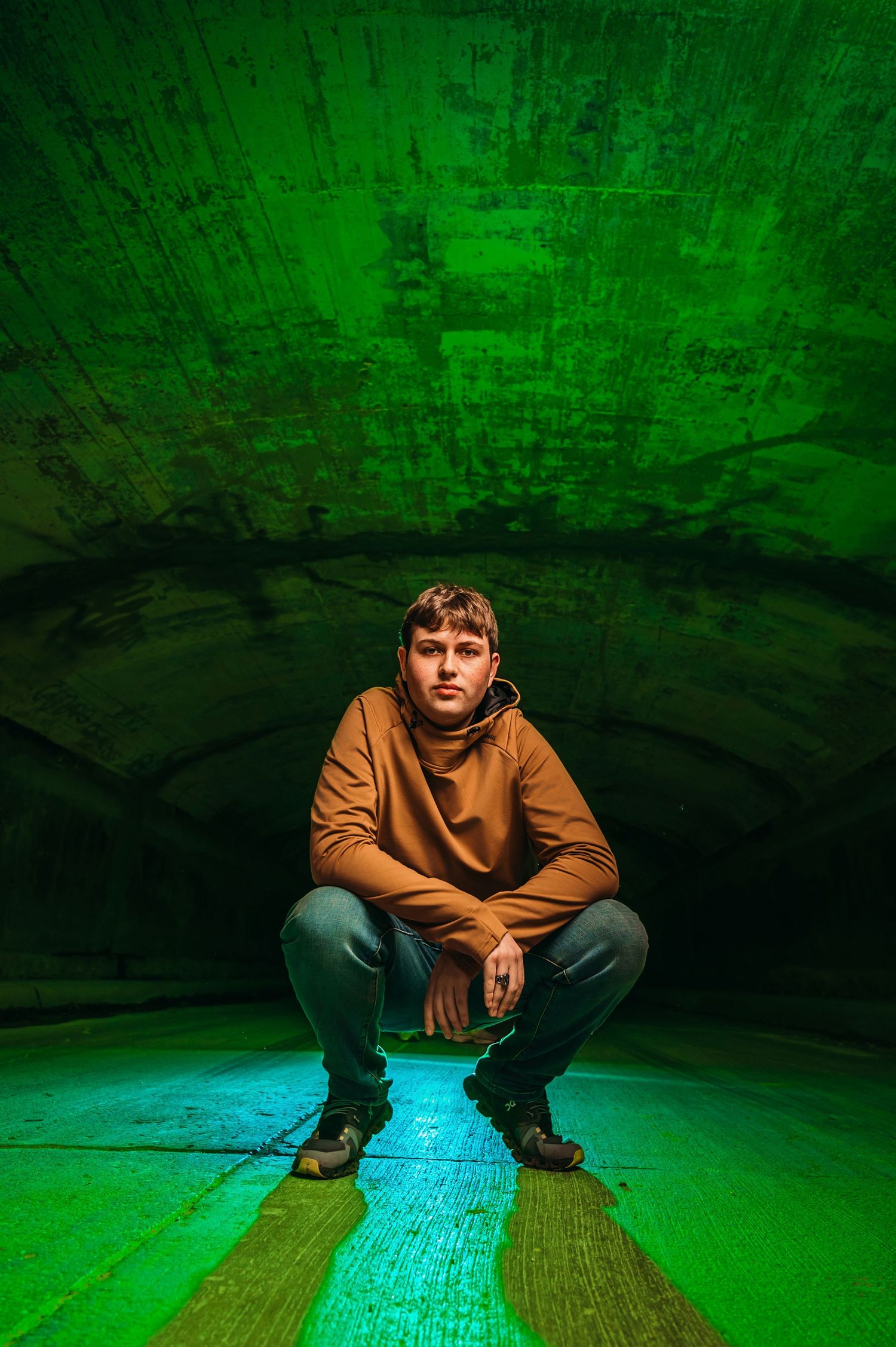 Young man in brown hoodie crouching in dramatic green-lit tunnel for creative portrait photography.