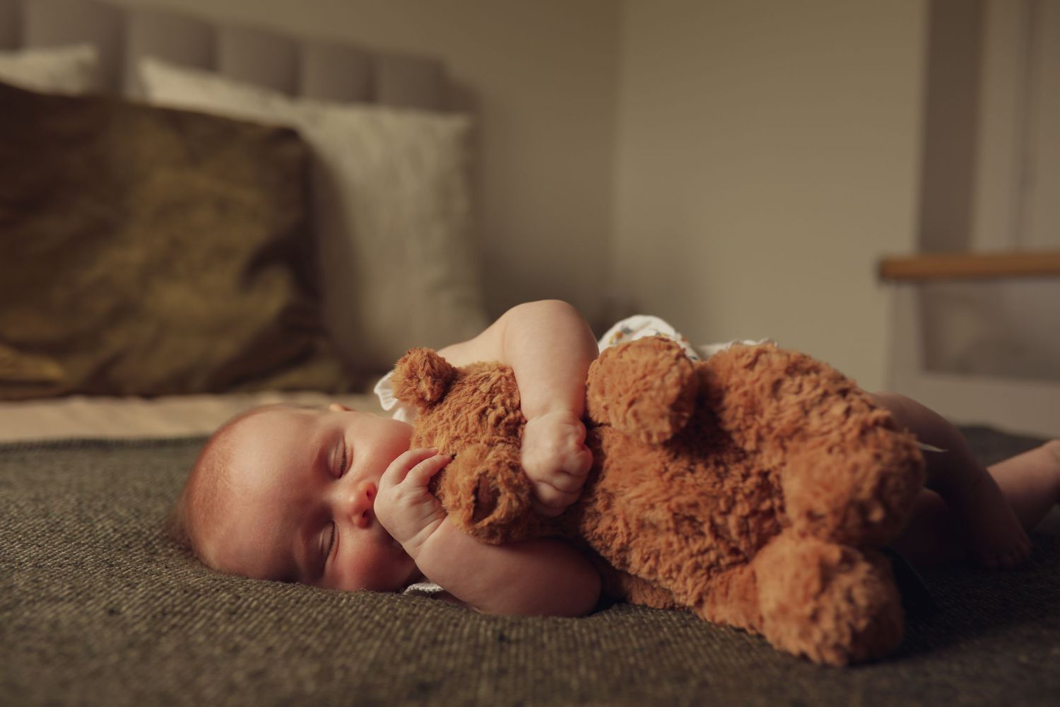 A sleeping baby cuddled up next to a large brown teddy bear on a gray couch.