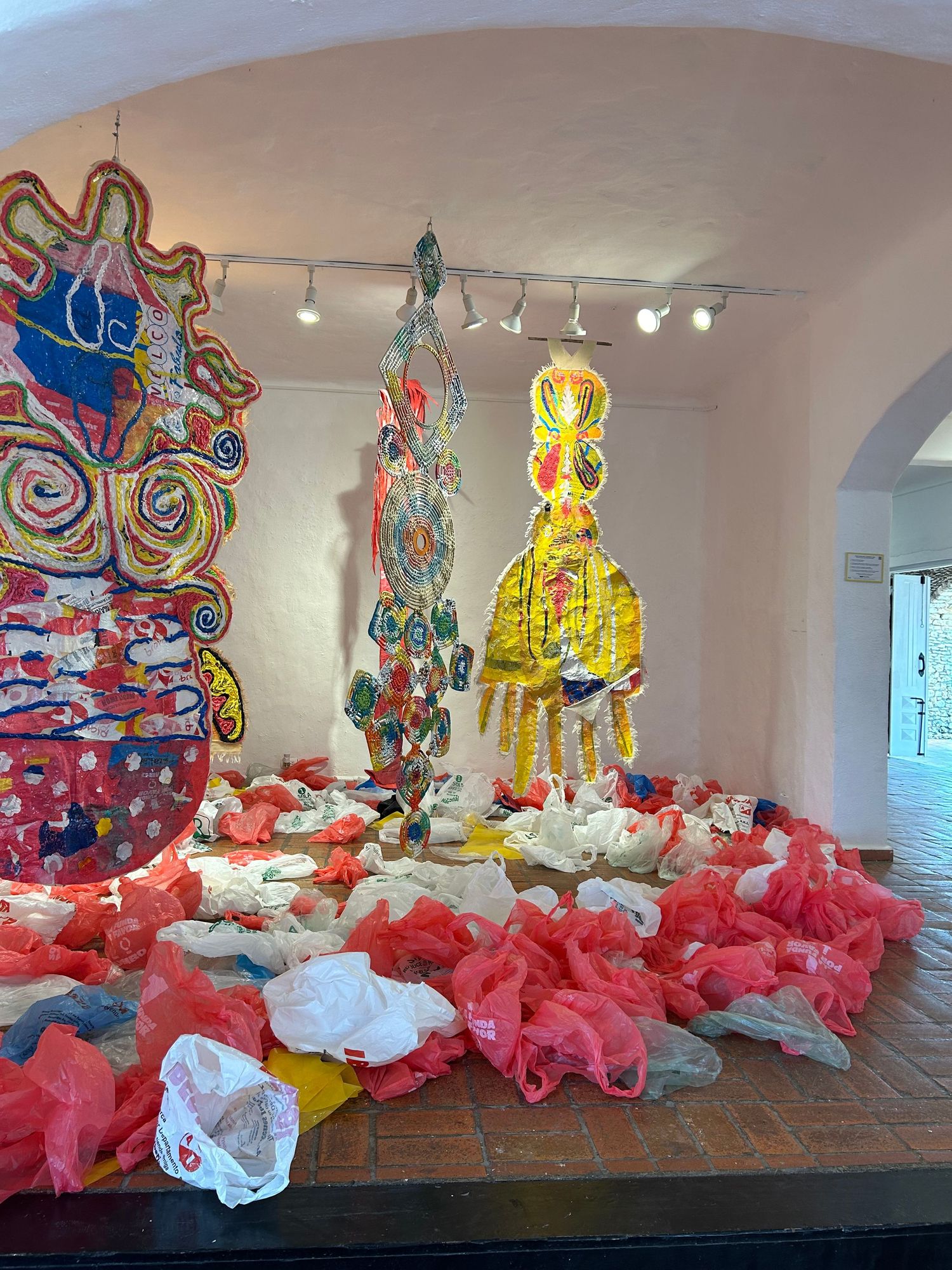 Vibrant gallery space featuring suspended plastic art pieces and red bags on tile floor.