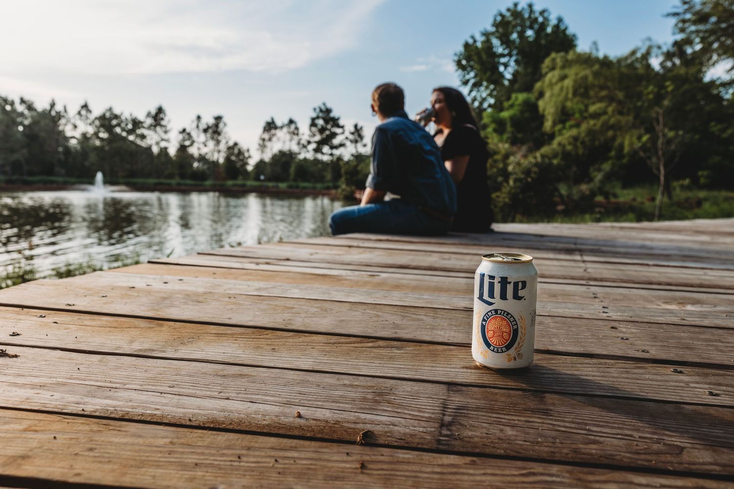 A Miller Lite can sits on wooden dock overlooking a serene lake with couple silhouetted in background.
