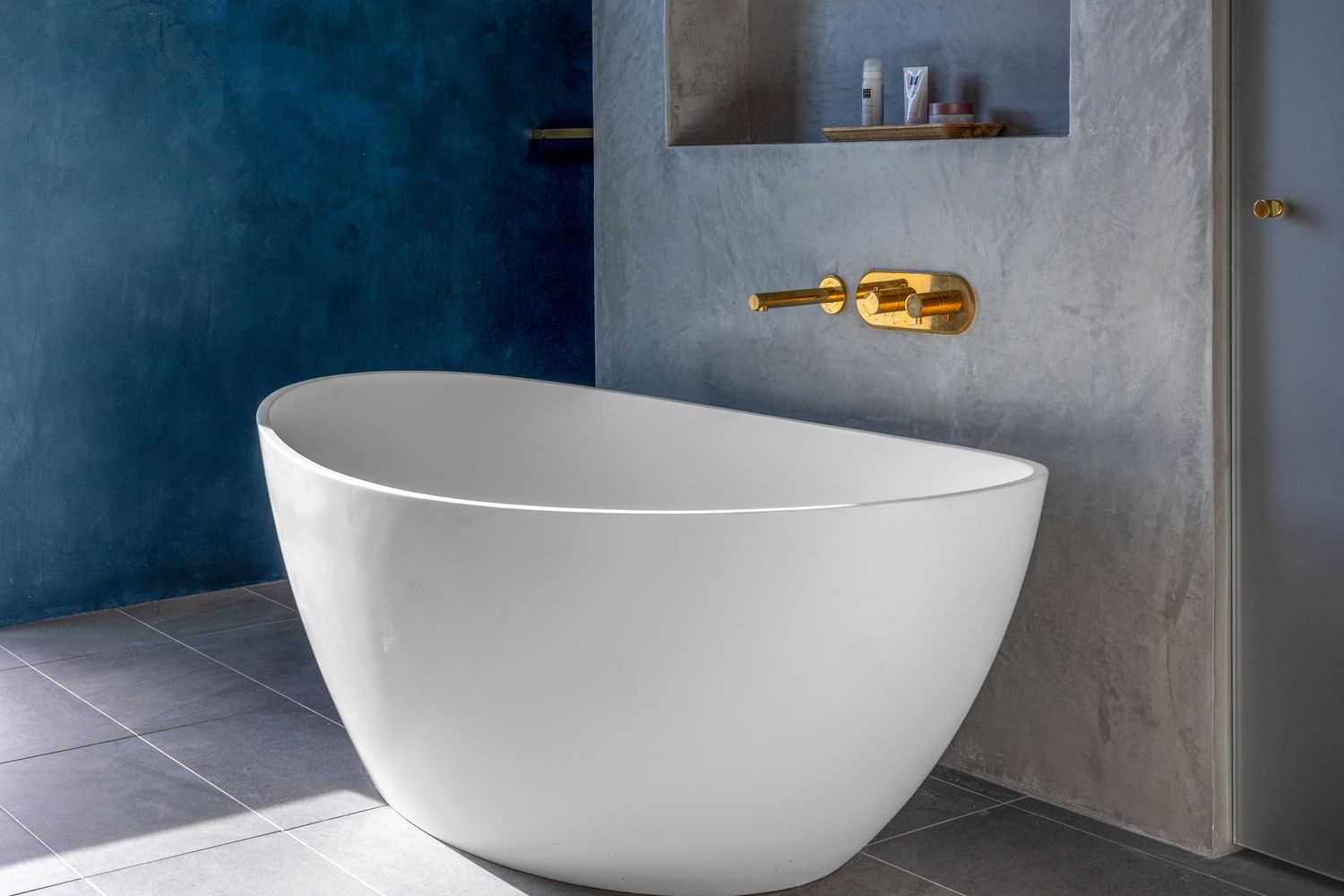 Modern freestanding white oval bathtub against blue and gray walls with gold wall-mounted faucet in luxury bathroom.