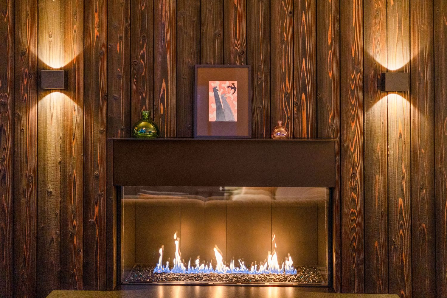 Modern gas fireplace with blue flames against dark wood paneled wall with ambient sconce lighting creates cozy atmosphere.