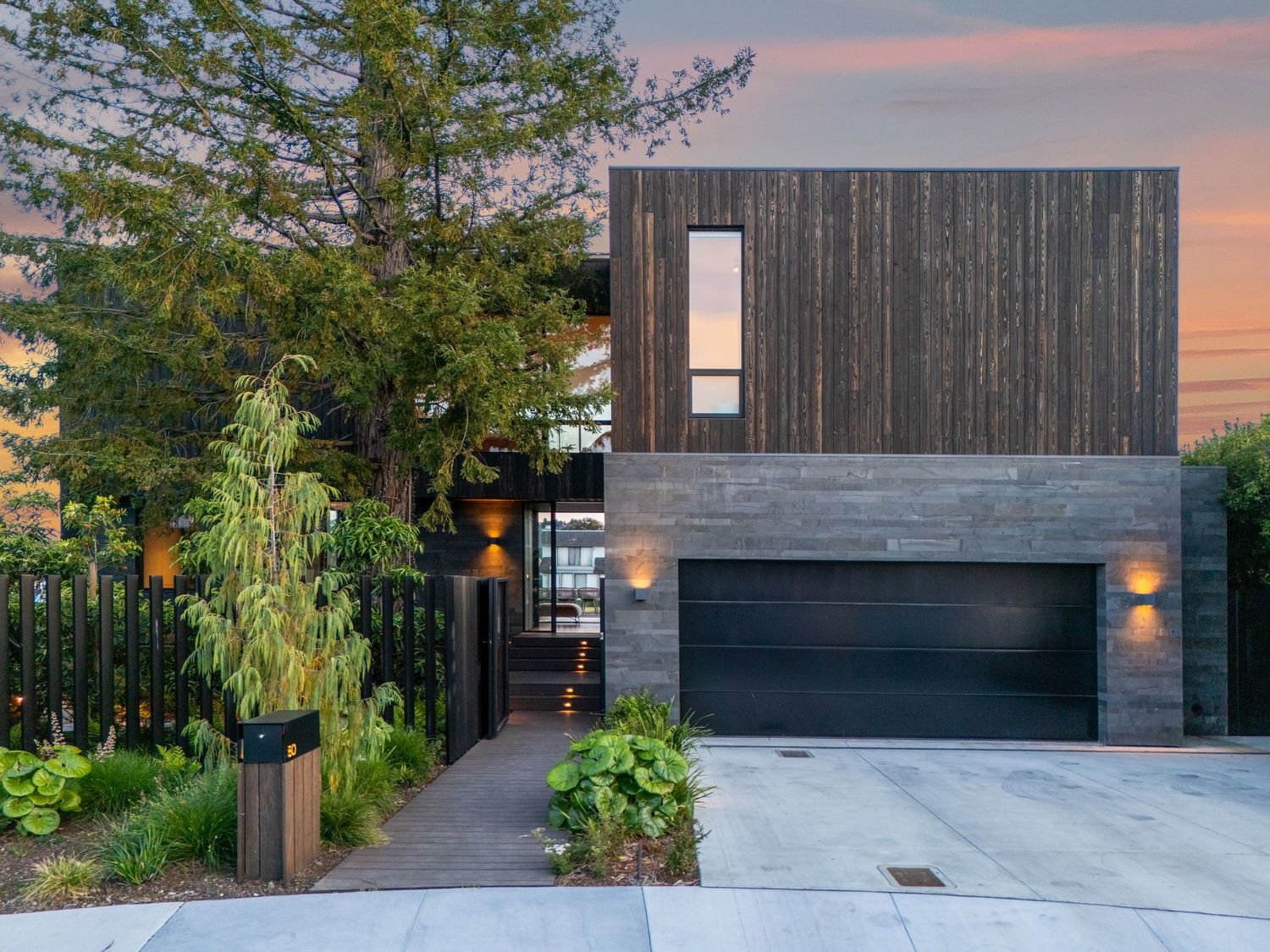 Modern two-story home with dark wood siding, concrete base, black garage door and lush landscaping at sunset.