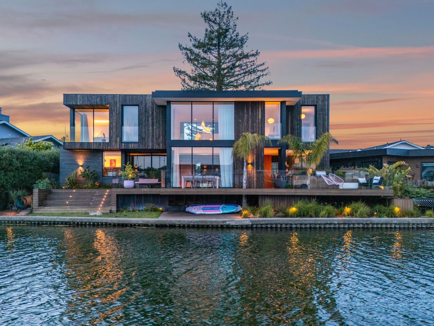Modern waterfront house with glass walls illuminated at sunset reflecting on calm water with paddle boards at the dock.