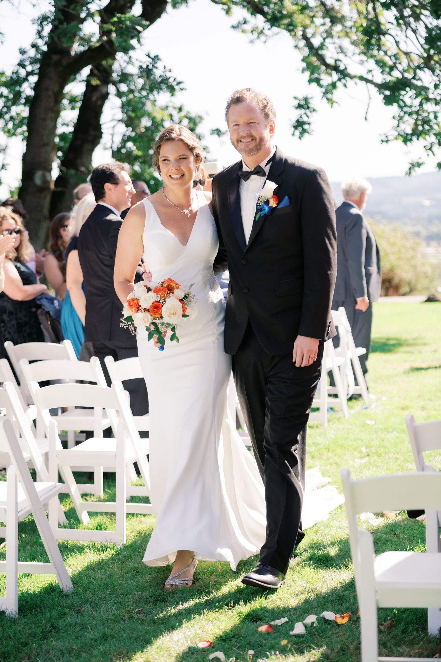 Wedding ceremony outdoors with white chairs and floral decorations on a sunny hillside.