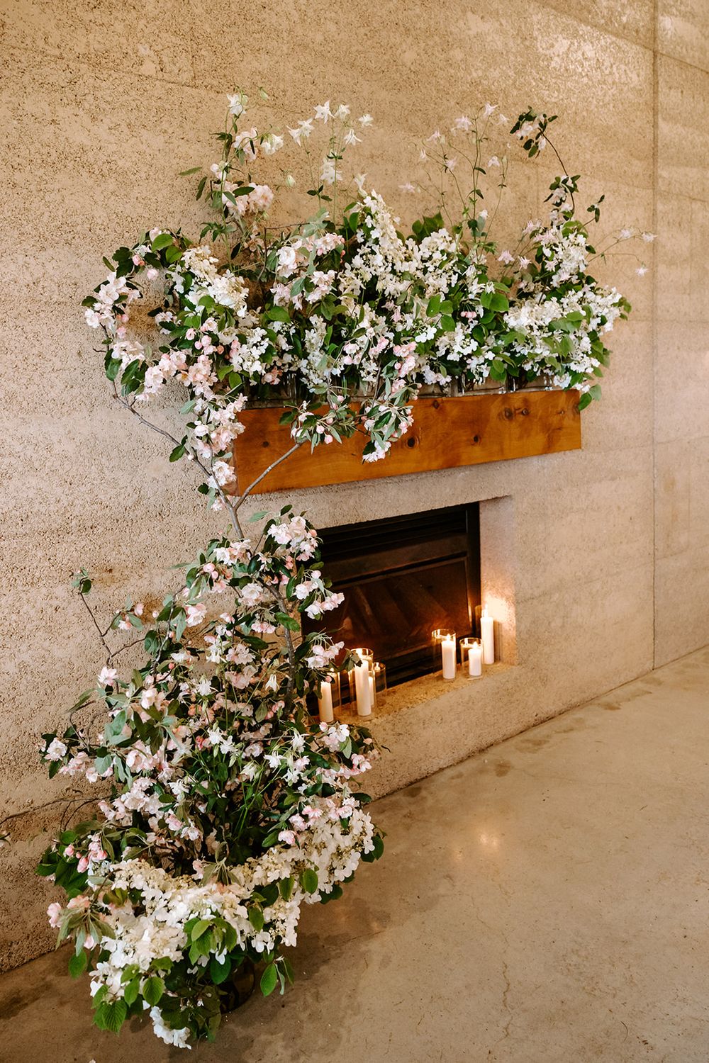Elegant white floral arrangements with cascading blooms decorate a stone fireplace with glowing candles.