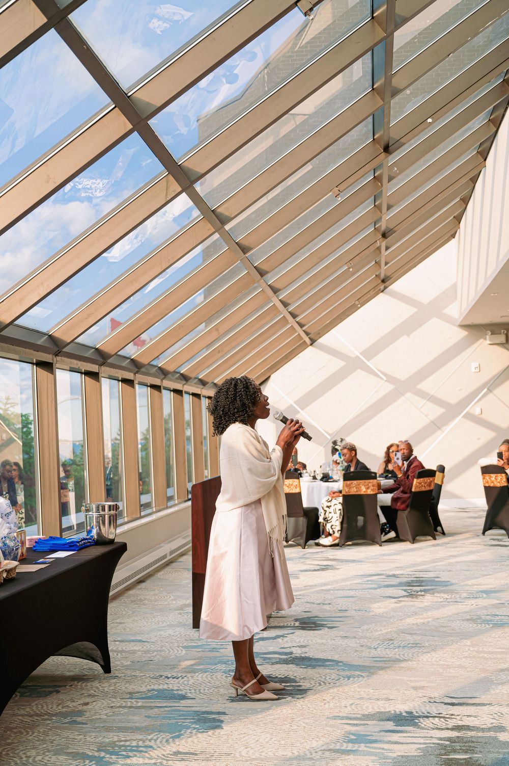 A performer in pink dress singing during an event in a glass-roofed venue.