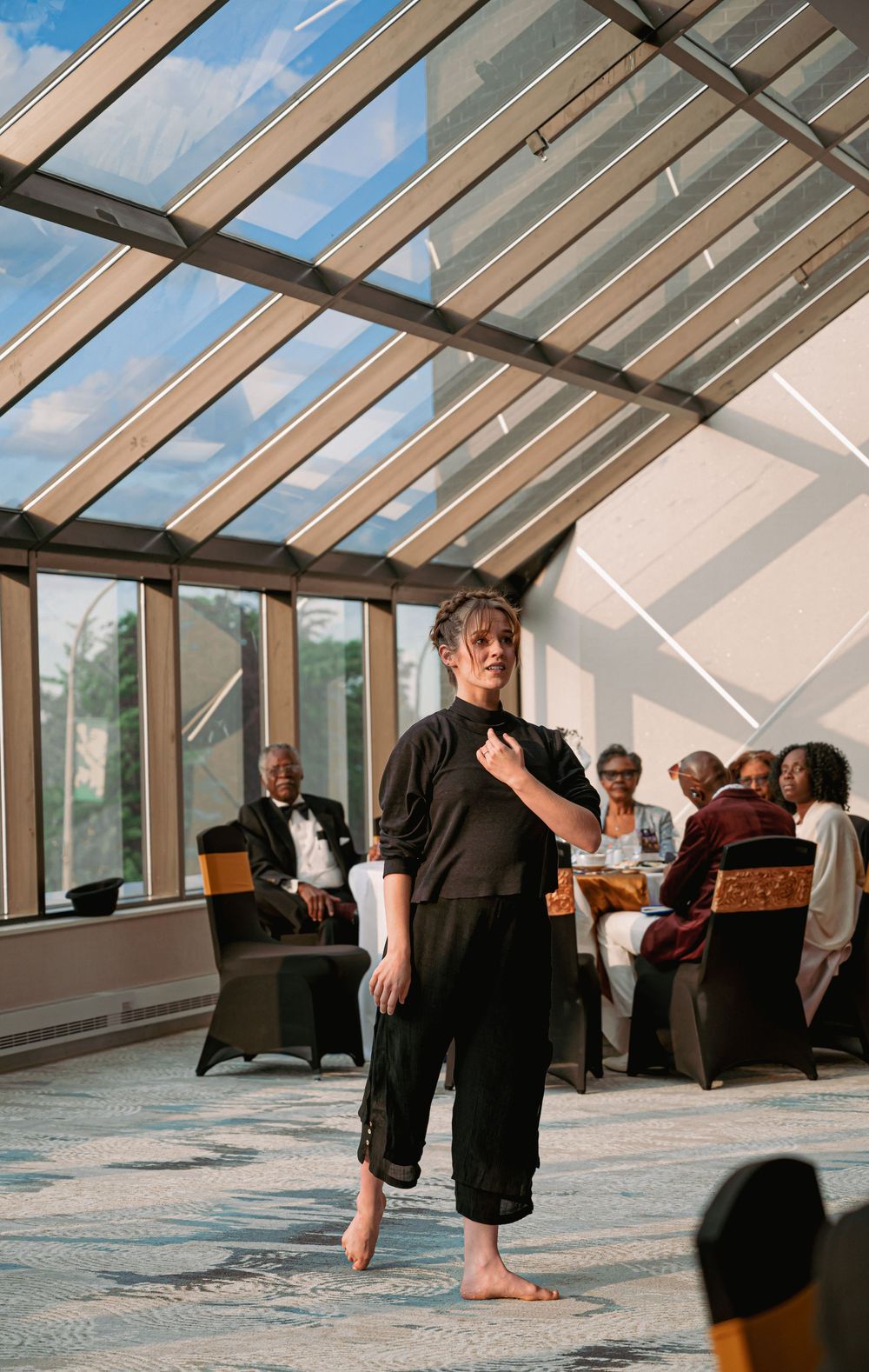 A performer in black attire presenting in a sunlit room with glass ceiling.