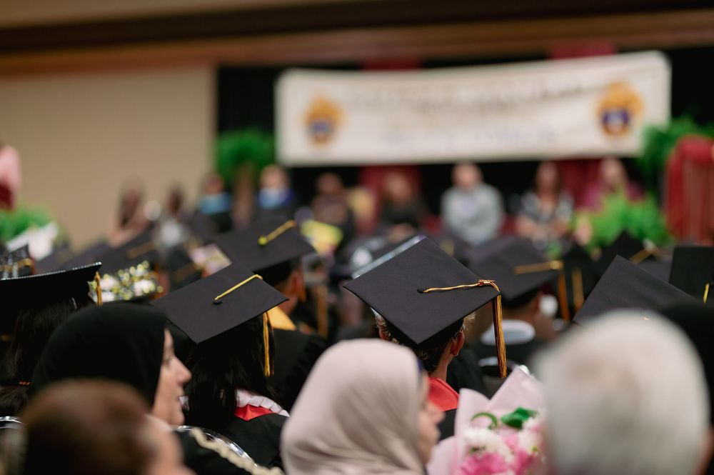 Graduation ceremony audience with mortarboards and hijabs visible in the crowd.