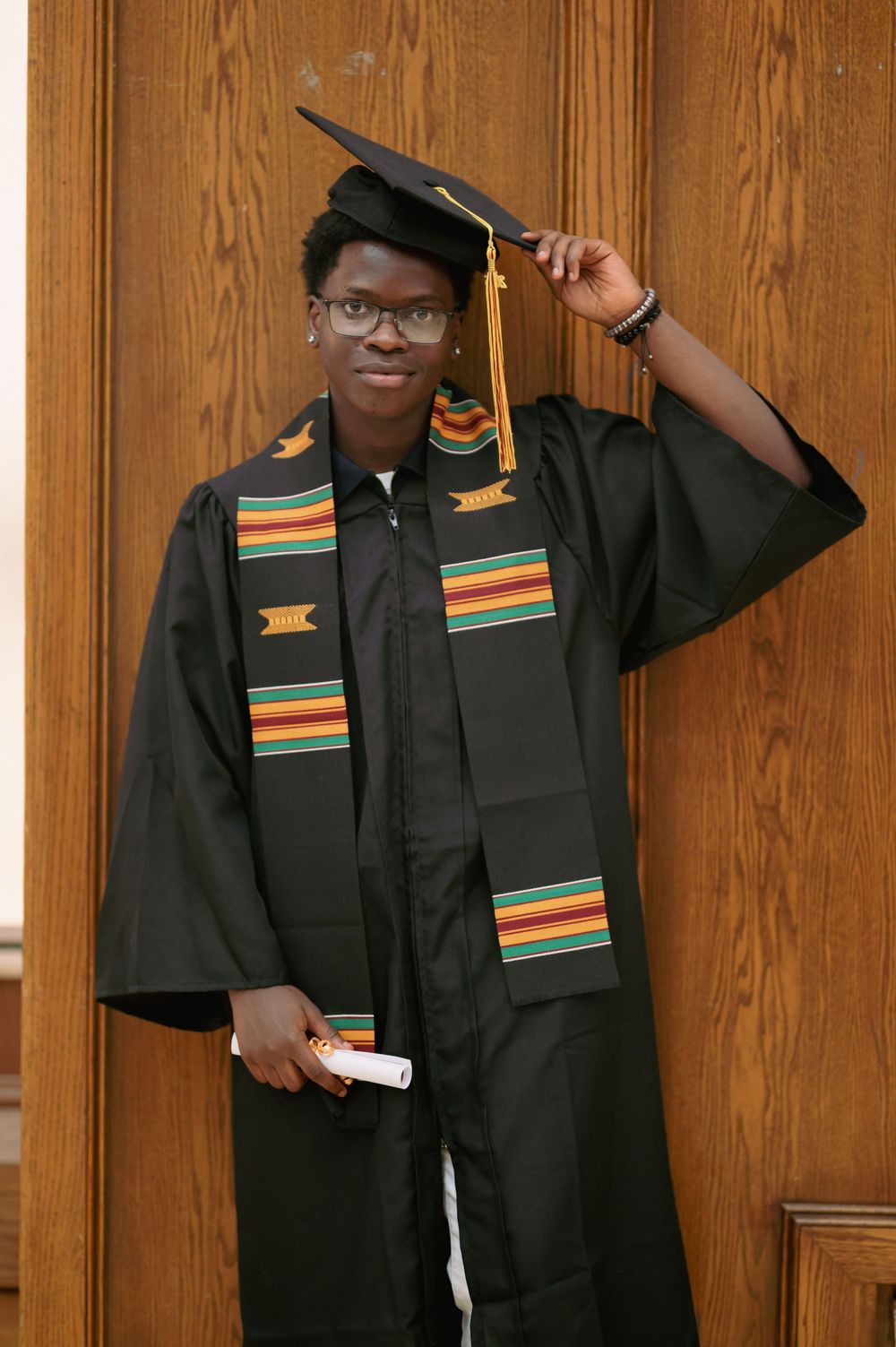 Graduate in traditional cap and gown with colorful kente stole against wooden backdrop.