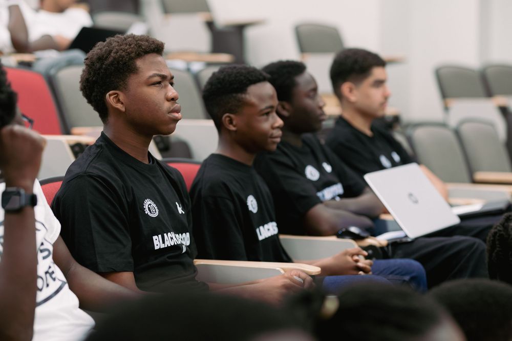 Students in matching black t-shirts seated in an auditorium during a presentation.