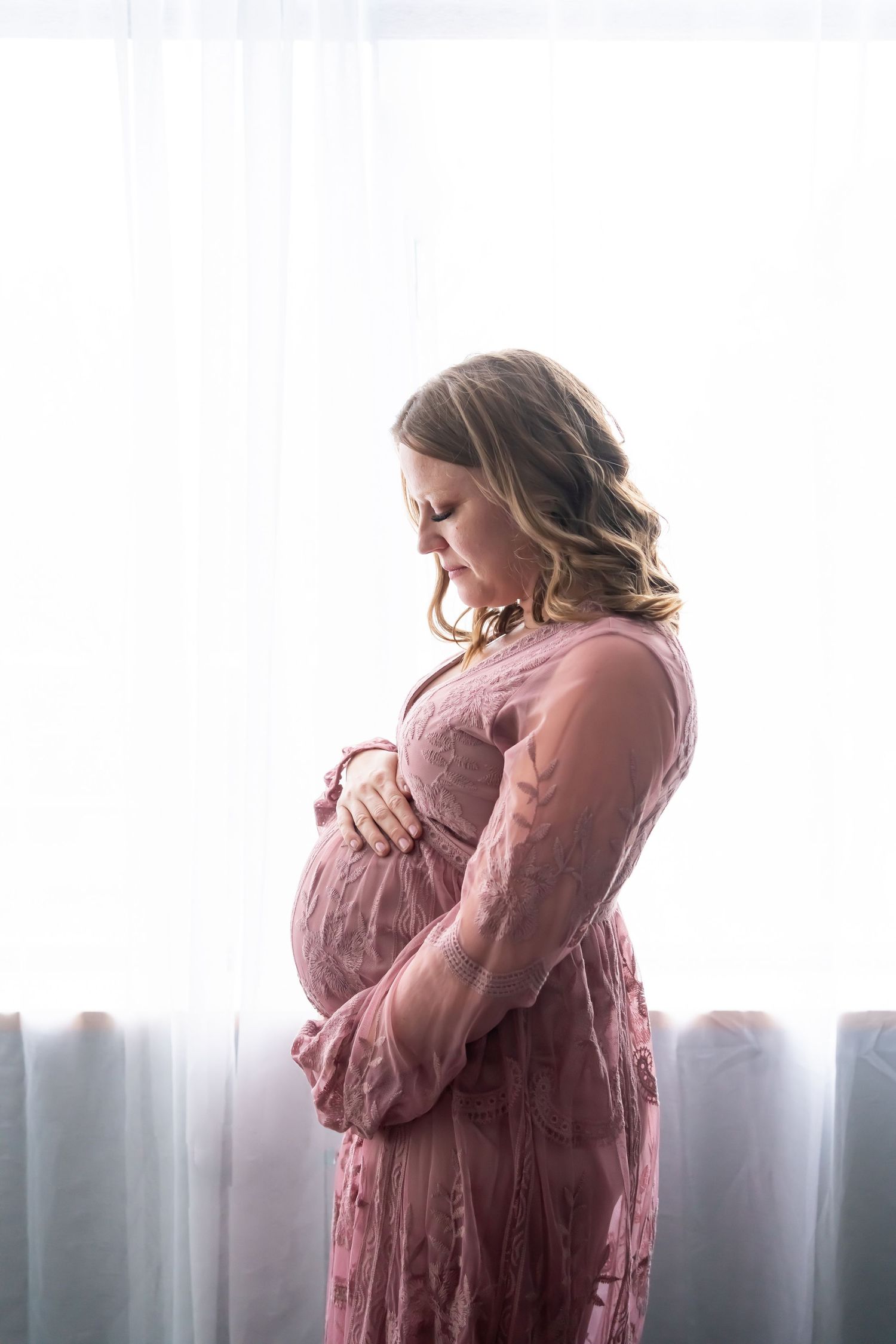 A silhouetted maternity portrait by window light in a pink vintage-style dress.