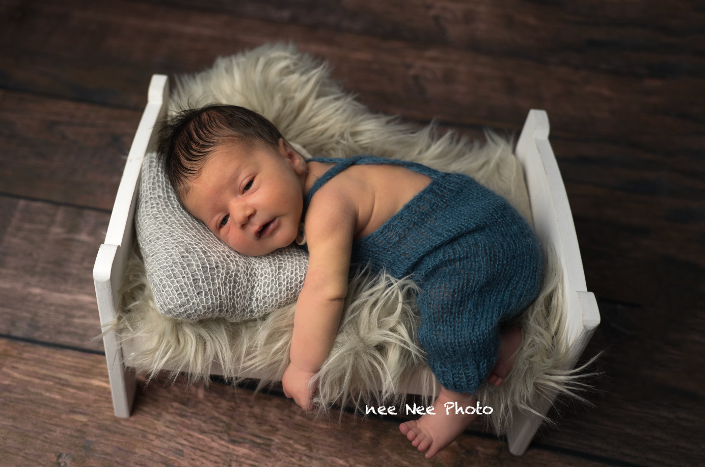 Newborn baby sleeping peacefully on fluffy fur in a tiny white bed, wearing a blue knit outfit, in a professional photo. Embracing Life’s Be