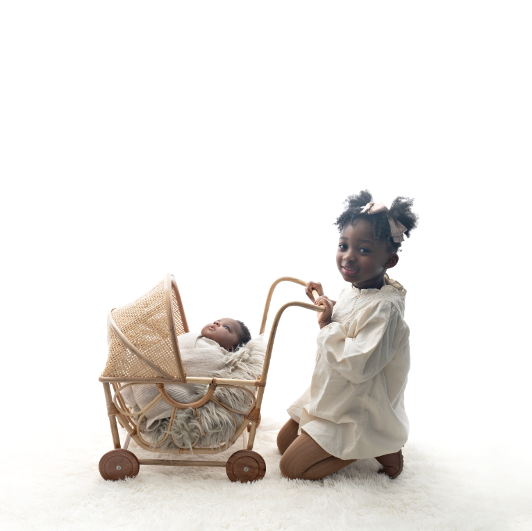 Young girl kneeling beside a wicker pram with a sleeping newborn baby wrapped in a beige blanket on a white background.