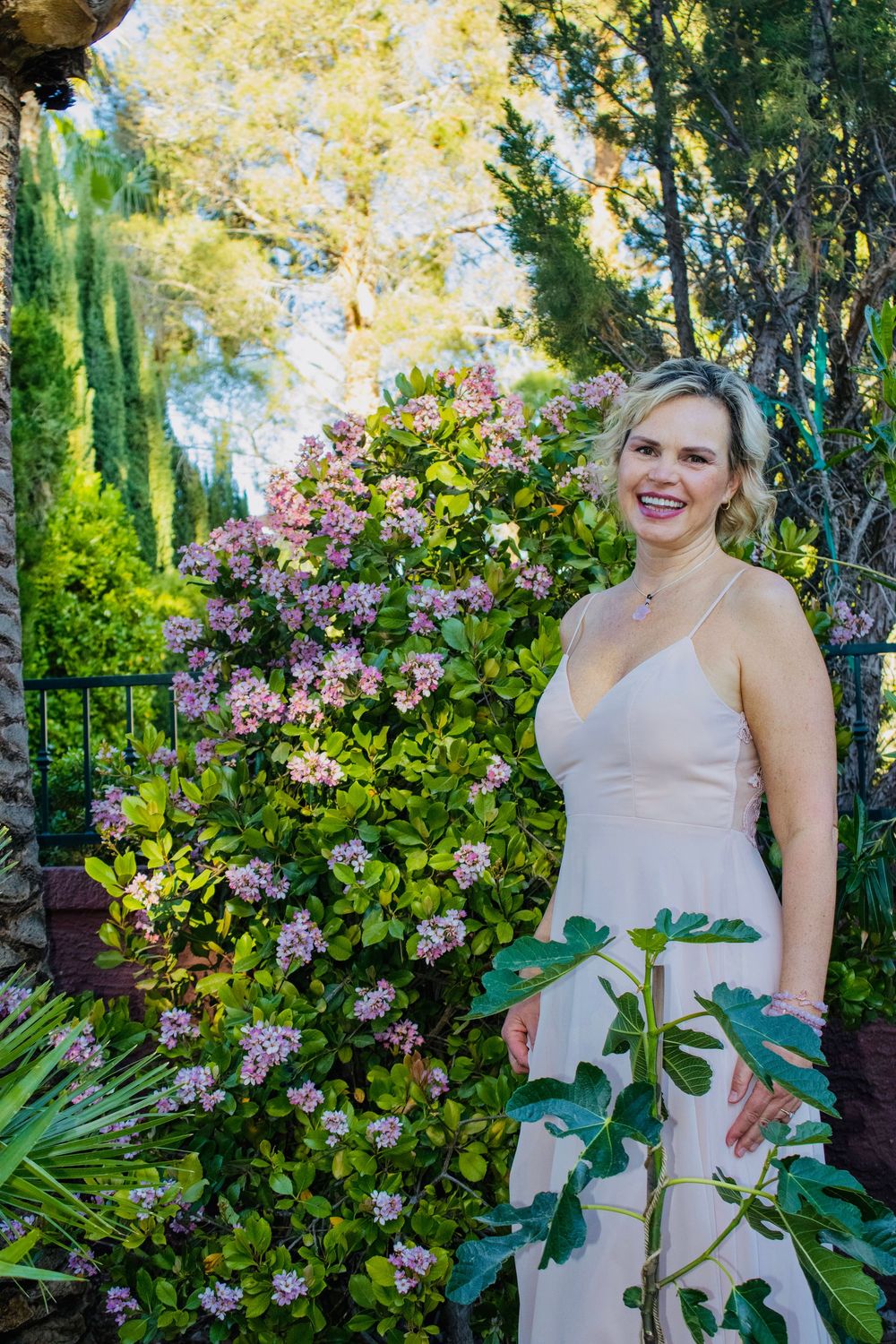 Smiling blonde woman in a blush spaghetti strap dress posing beside blooming pink flowers in a lush garden.
