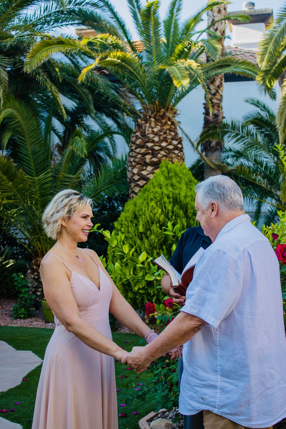 Smiling bride in blush pink dress holds hands with groom during outdoor wedding ceremony surrounded by tropical palm trees.