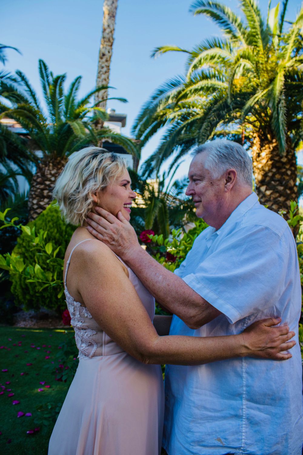 Older couple sharing a romantic moment outdoors, surrounded by lush tropical palm trees and colorful flowers.