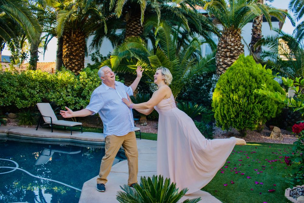 Couple dancing joyfully by a pool surrounded by lush palm trees in a sunny tropical garden setting.