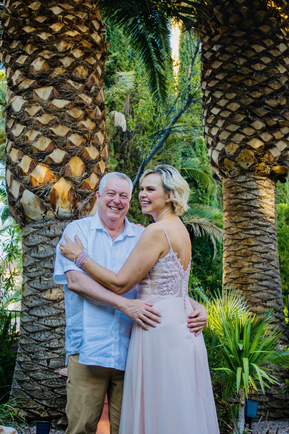 Smiling couple embracing among tall palm trees in a tropical garden, woman wearing elegant backless blush gown.
