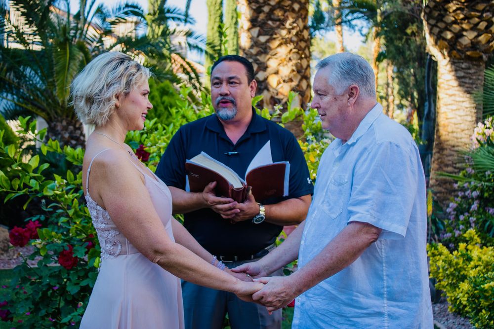Couple exchanging vows outdoors with officiant holding book, surrounded by tropical palms and red roses.