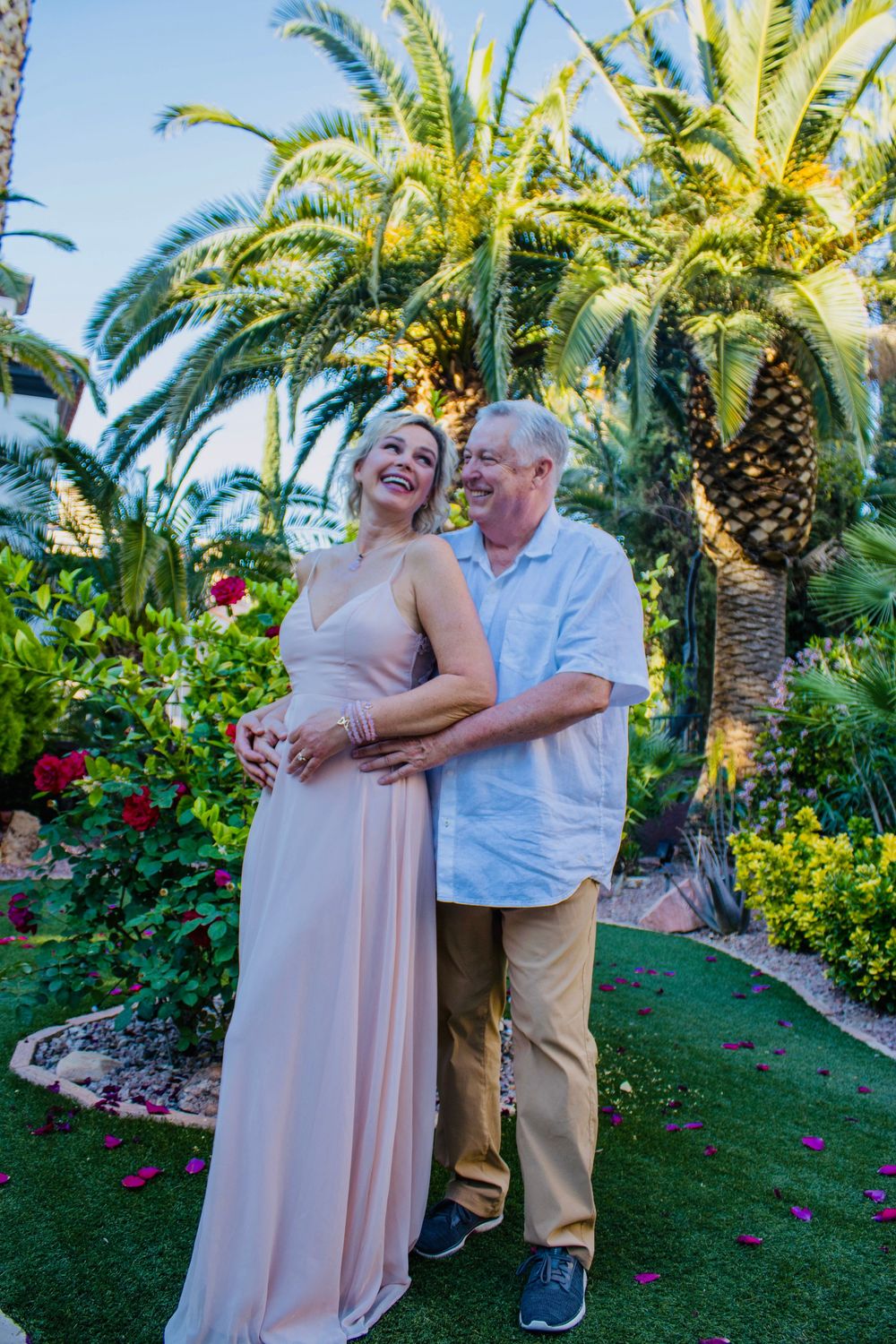 Smiling couple embraces in tropical garden, woman in pink gown and man in white shirt among palm trees and red roses.
