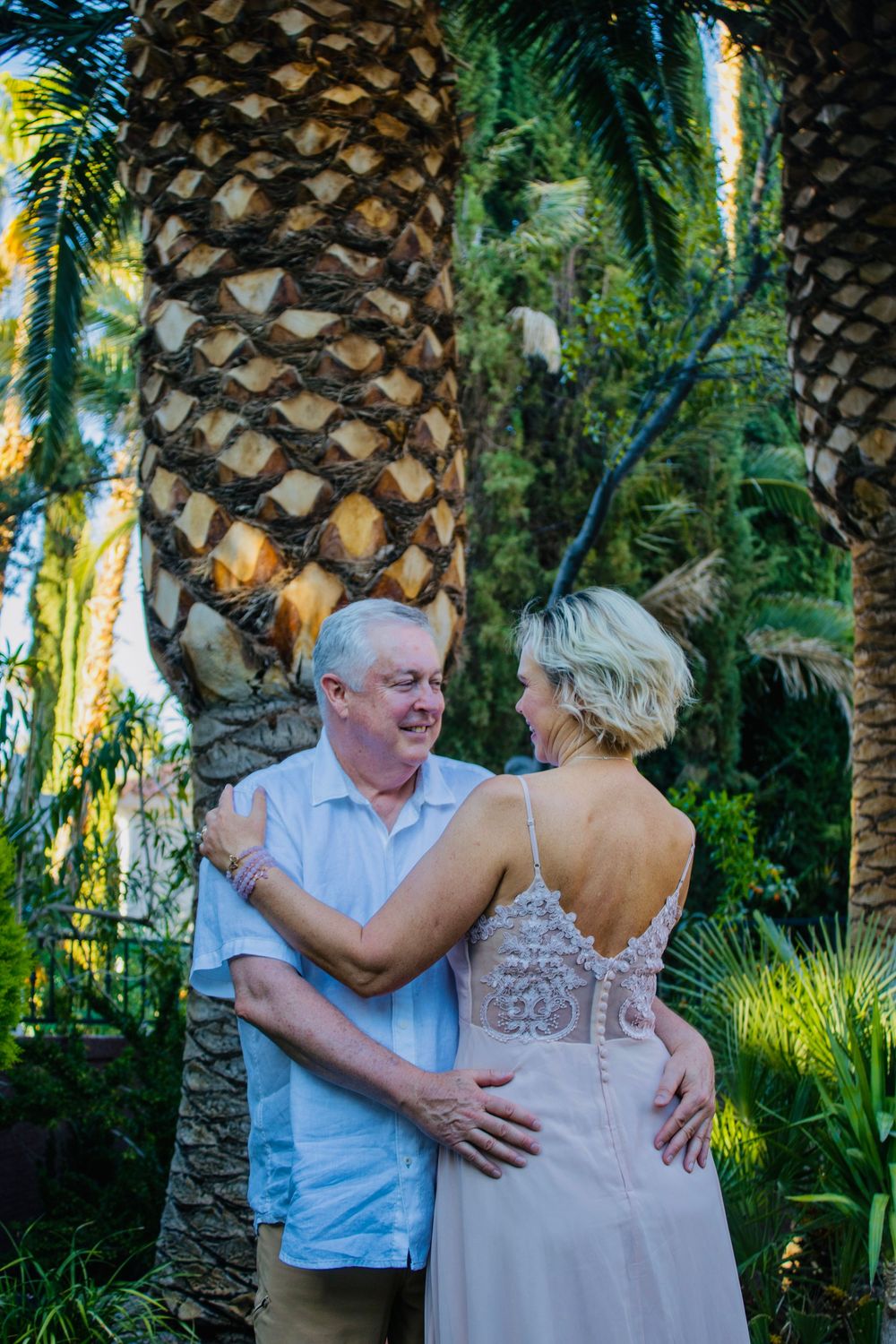 Couple sharing a tender moment among lush palm trees, bride in backless lace gown and groom in light blue shirt.