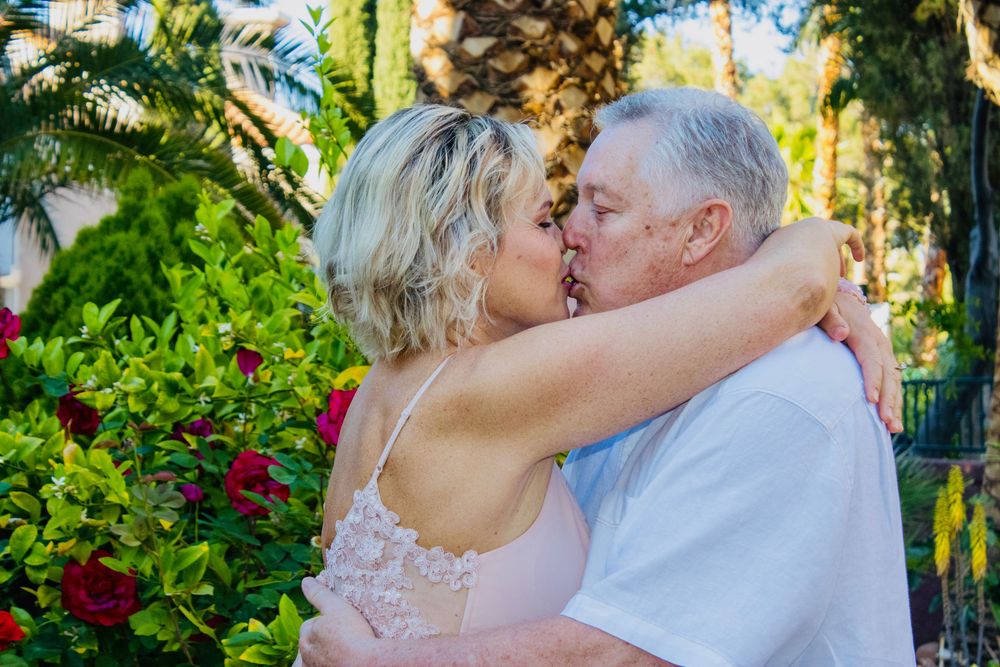Older couple sharing a romantic kiss outdoors surrounded by lush greenery and red roses.